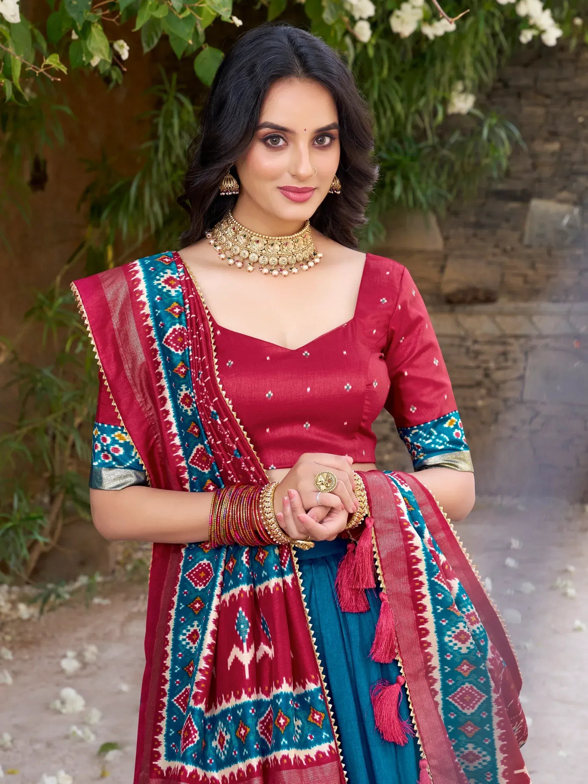 Woman in red and teal ethnic lehenga choli with intricate patterns and gold jewelry, perfect for festive occasions.