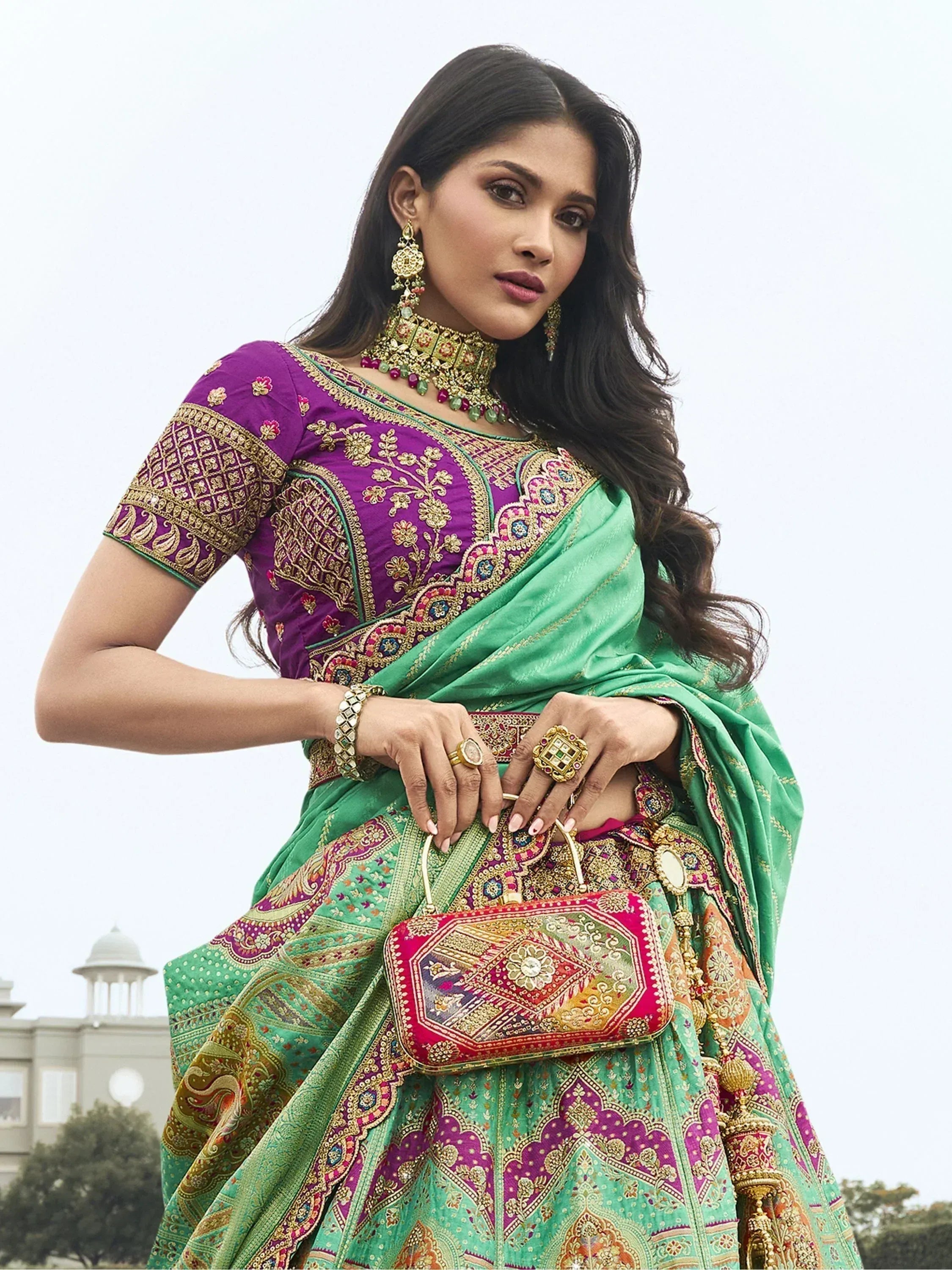 Woman in vibrant purple and green embroidered lehenga choli with traditional jewelry and clutch, perfect for Indian ethnic wear occasions.