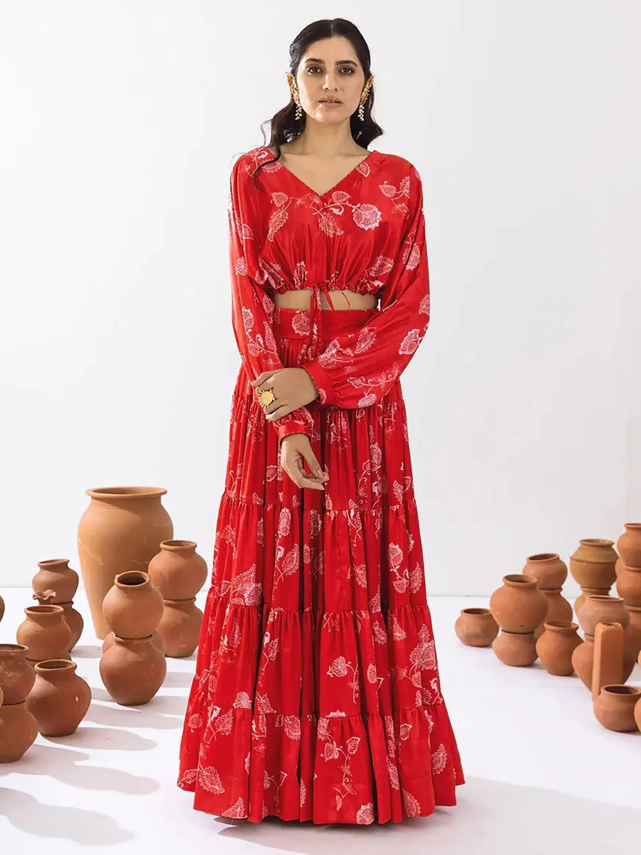 Red floral print ethnic gown with tiered skirt and long sleeves, styled with gold jewelry and clay pots