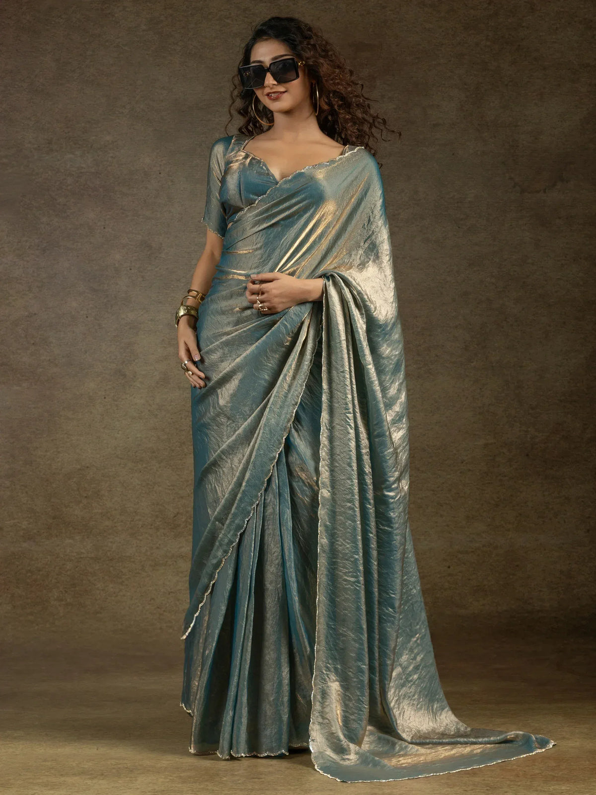 Woman in elegant teal and gold metallic saree with scalloped border, styled with sunglasses and gold jewelry.