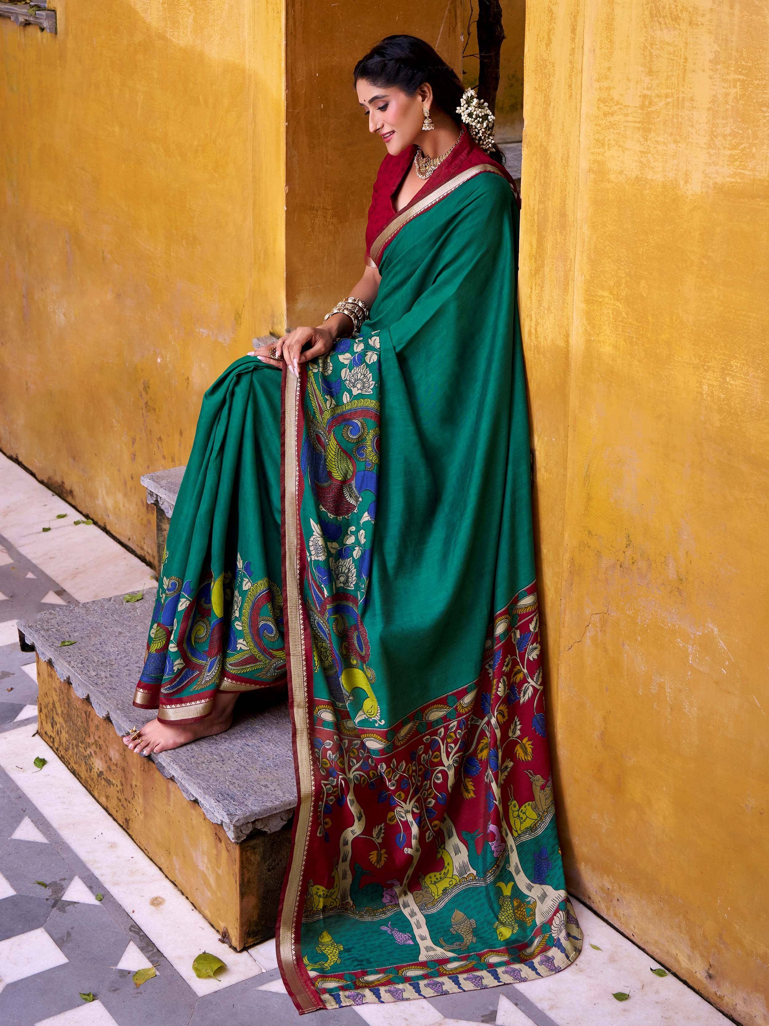 Woman in green and red printed saree with gold border, sitting on steps against yellow wall