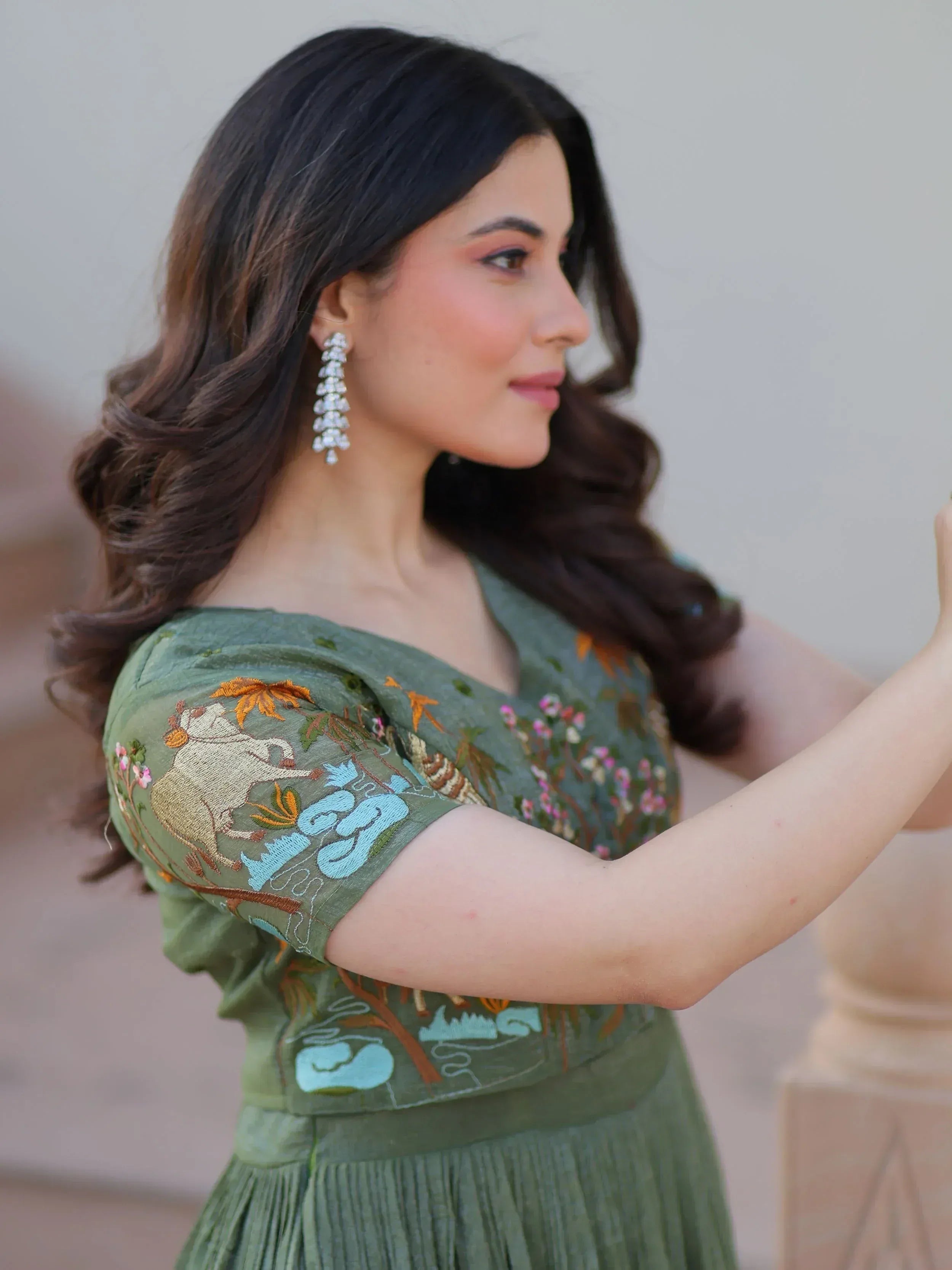Woman in olive green embroidered ethnic gown with floral and animal motifs, paired with dangling earrings.