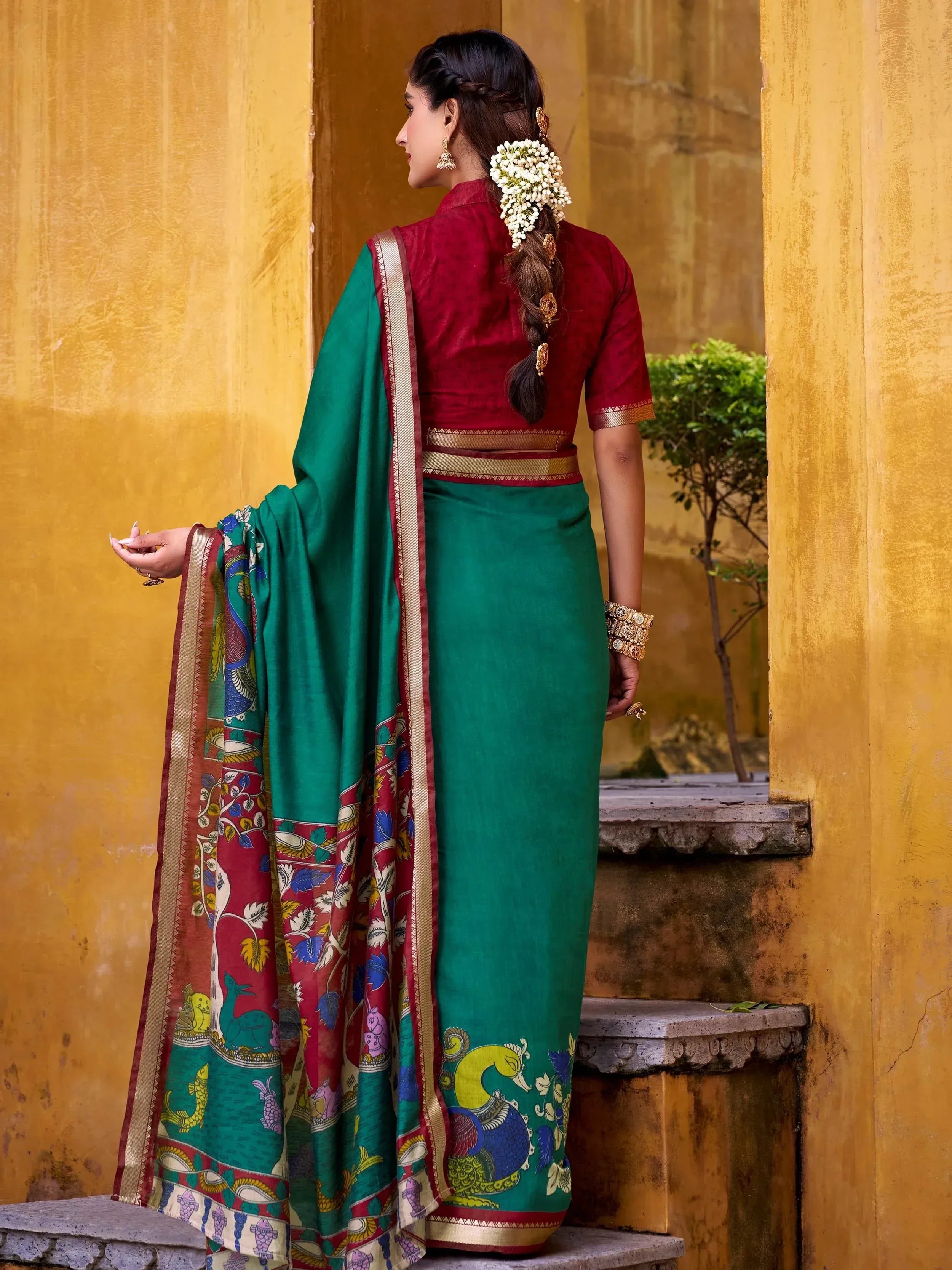 Woman in green printed saree with red blouse, floral hair accessory, standing by yellow wall