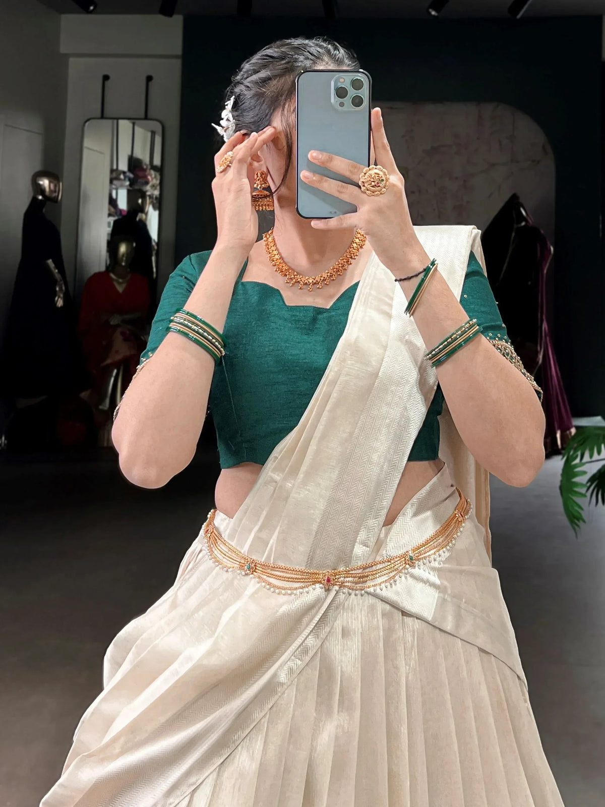 Woman in white saree with green blouse, gold jewelry, and bangles, taking mirror selfie in boutique