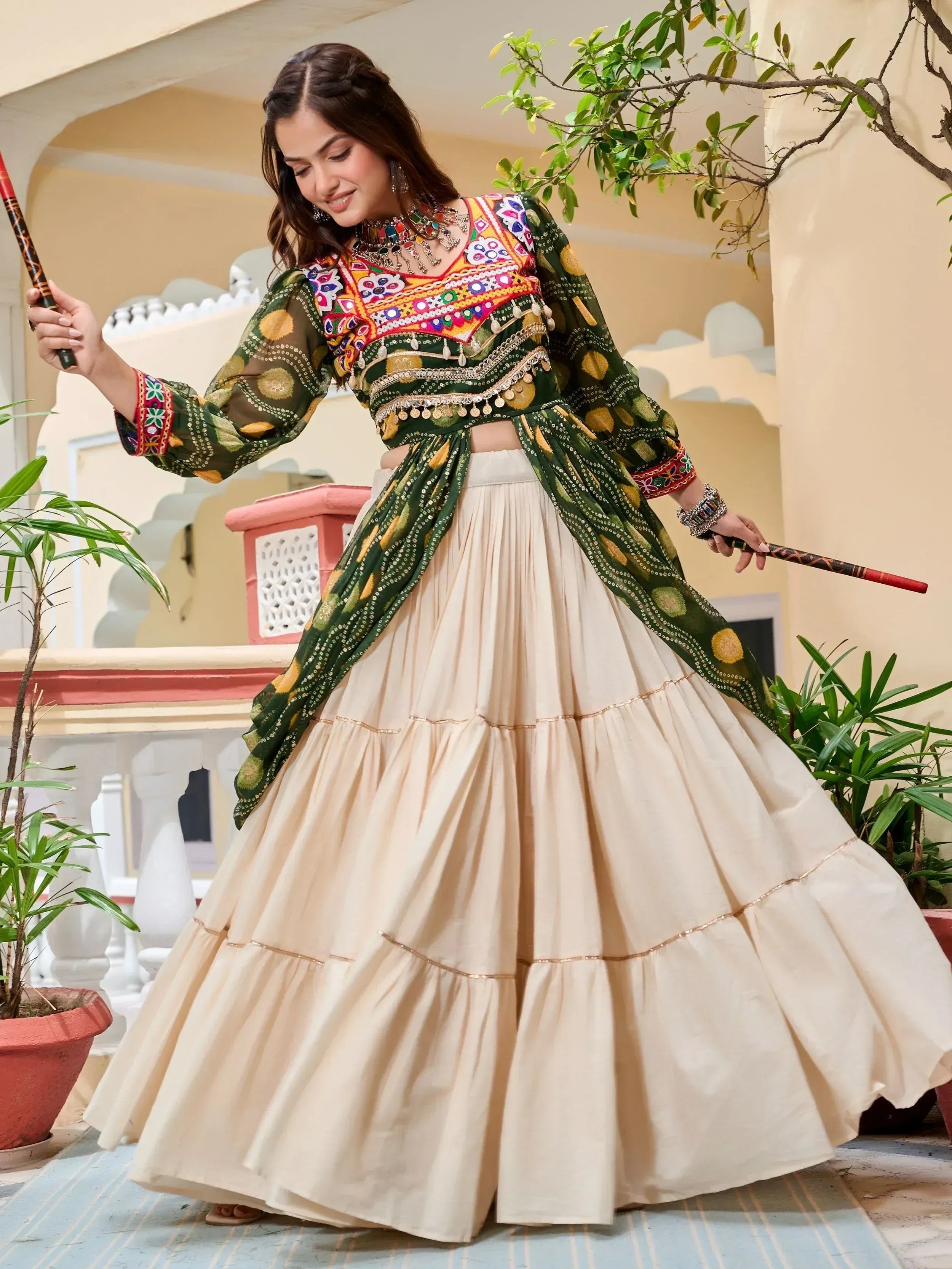 Woman in vibrant green embroidered choli with cream tiered lehenga, holding dandiya sticks for festive Indian wear.