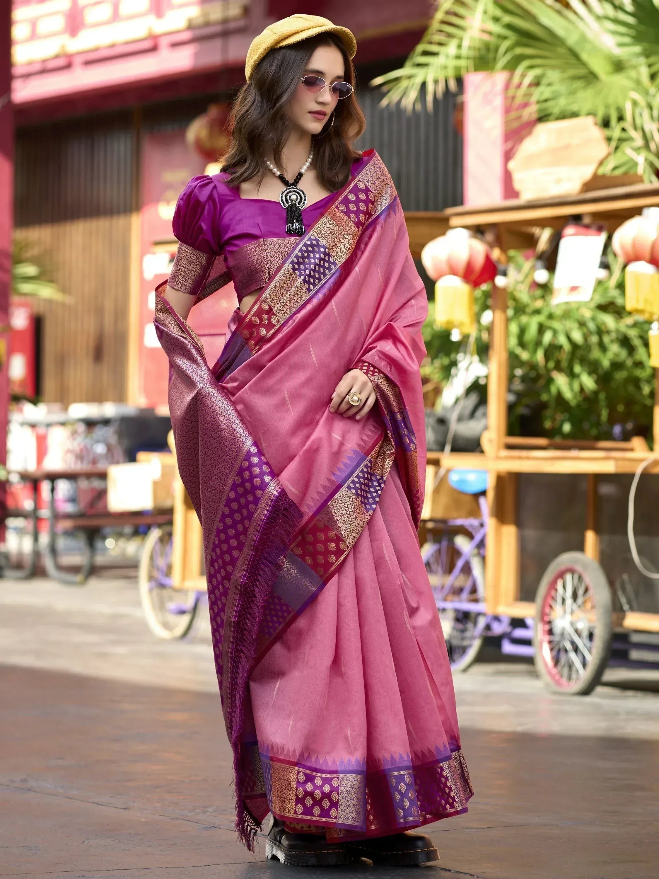 Woman in pink and purple designer saree with gold border, styled with yellow hat and sunglasses for ethnic fashion