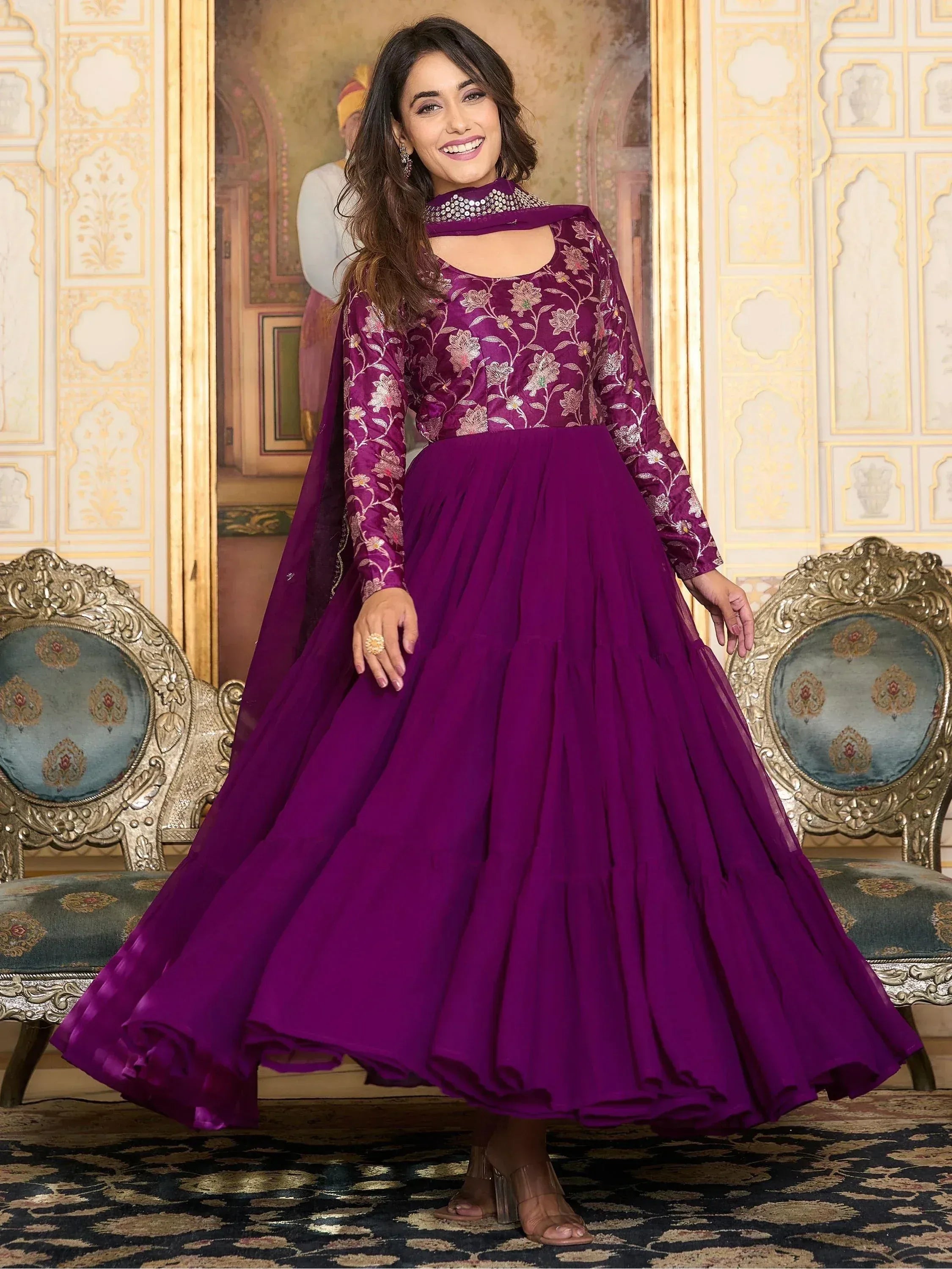 Woman in a purple embroidered ethnic gown with long sleeves, standing in a traditional setting.