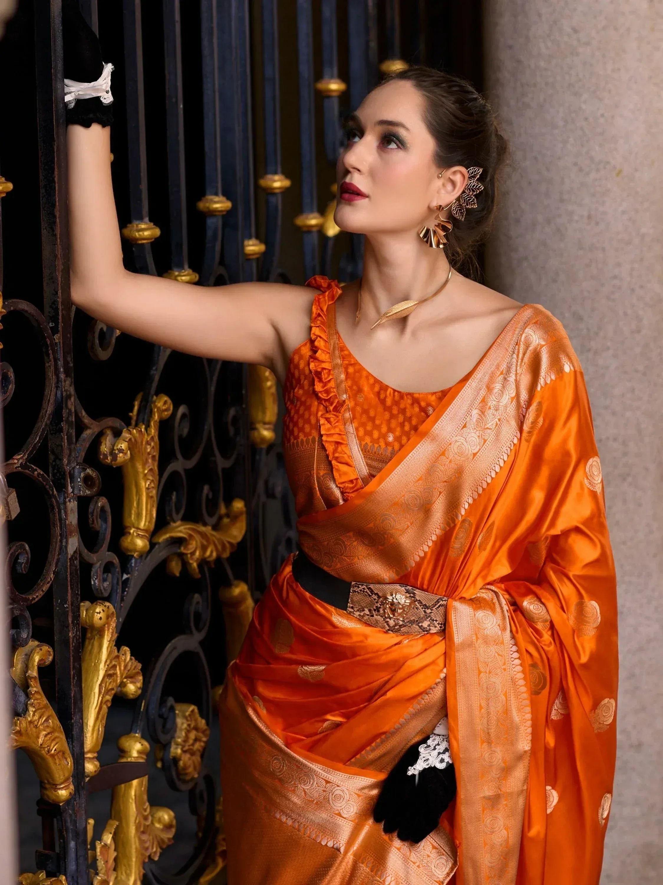 Woman in vibrant orange designer saree with gold motifs, black belt, and statement jewelry, posing by ornate gate