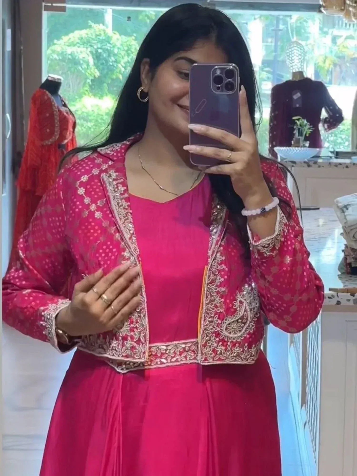 Woman in pink embroidered ethnic gown with matching jacket, taking mirror selfie in boutique