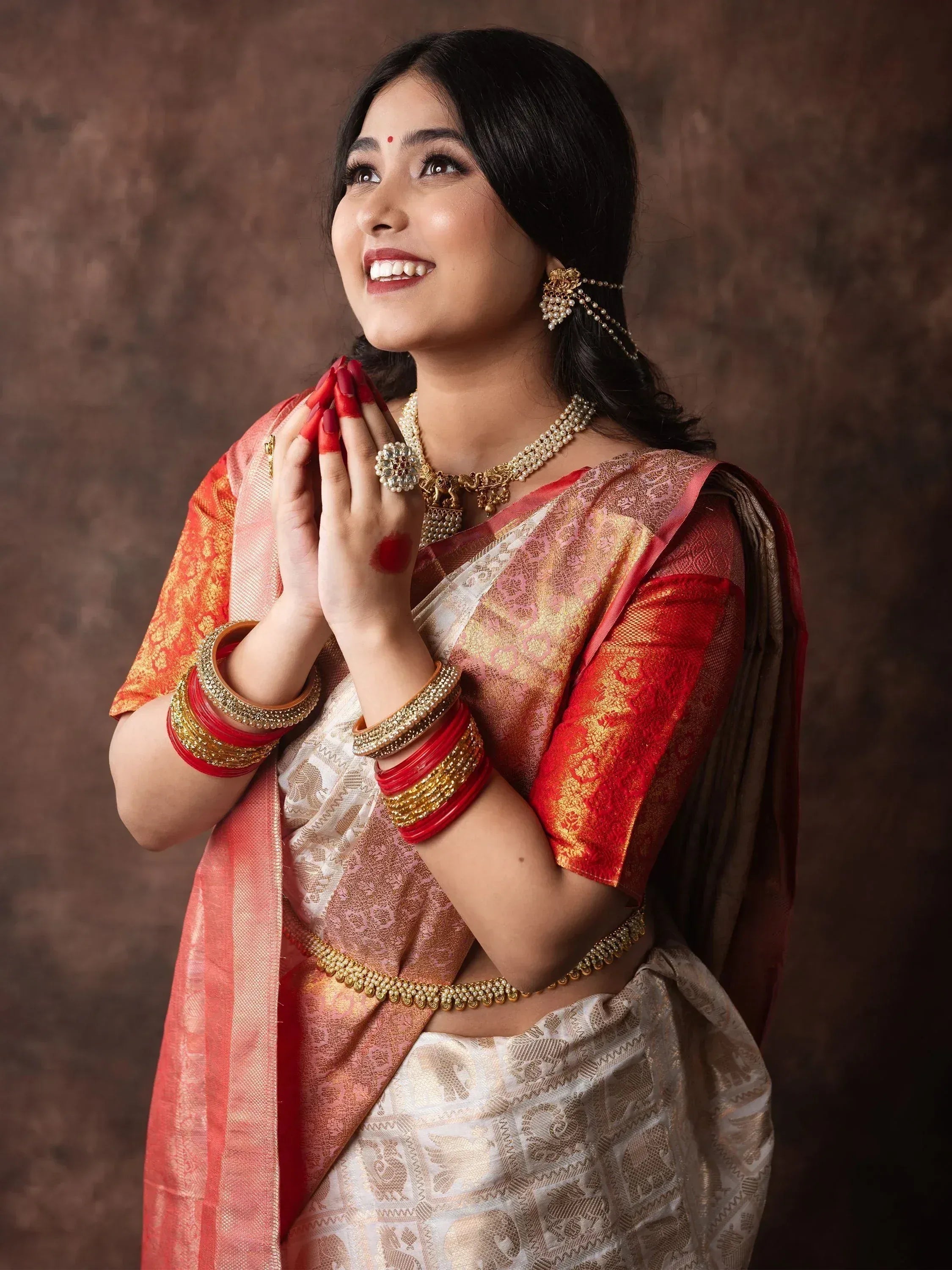 Woman in traditional Indian saree with gold jewelry, red bangles, and festive makeup, hands in prayer pose