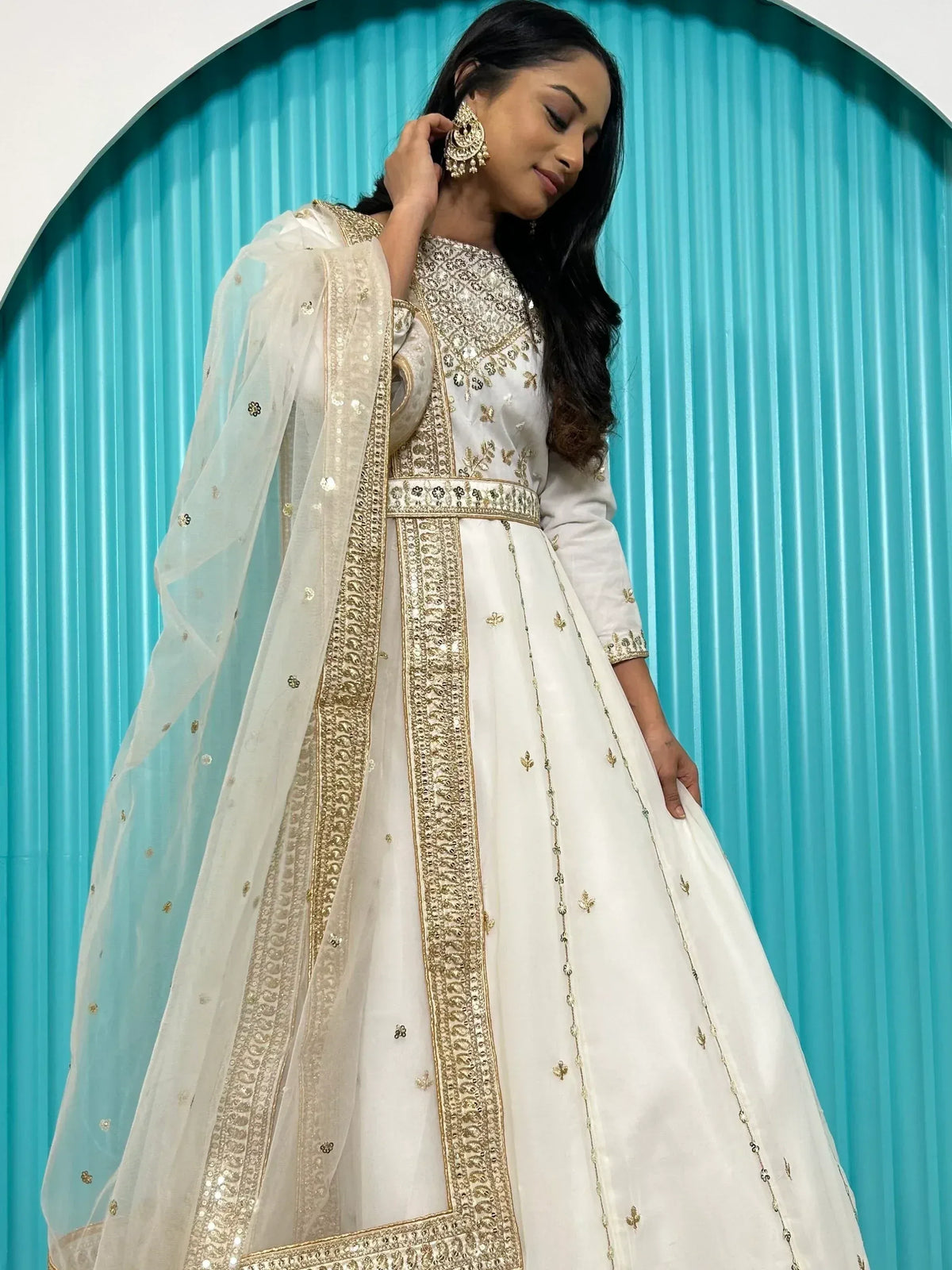 White embroidered anarkali with gold detailing, sheer dupatta, and statement earrings against teal backdrop