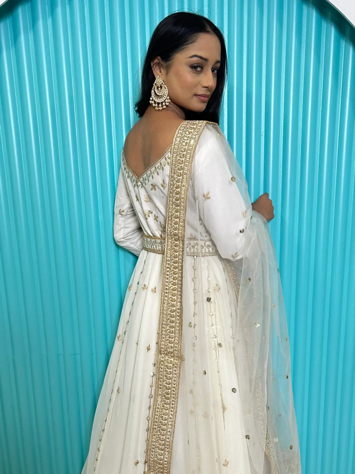 White embroidered anarkali with gold detailing, sheer dupatta, and statement earrings against blue backdrop