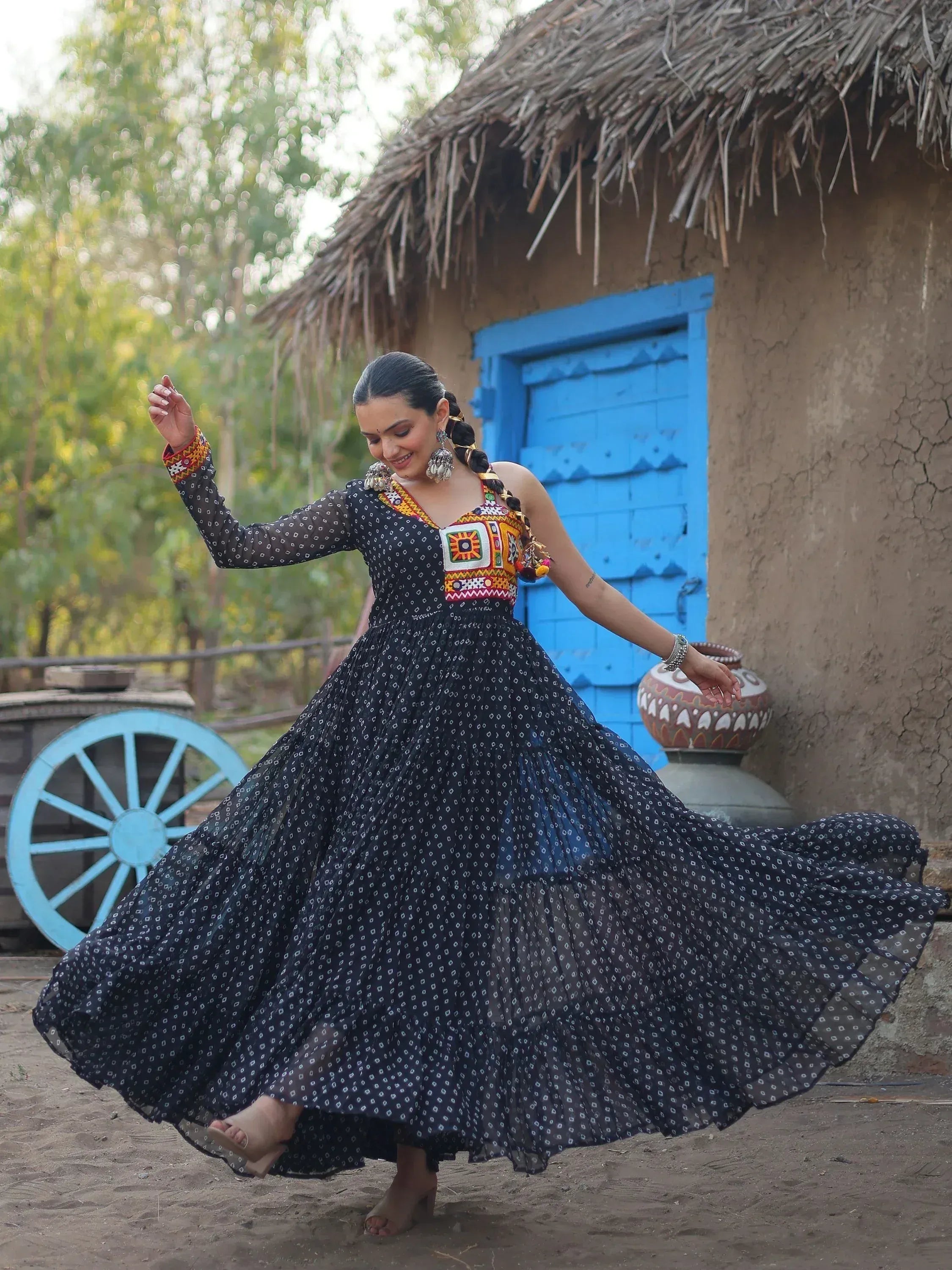Woman twirling in black printed ethnic gown with colorful embroidery, rustic hut background
