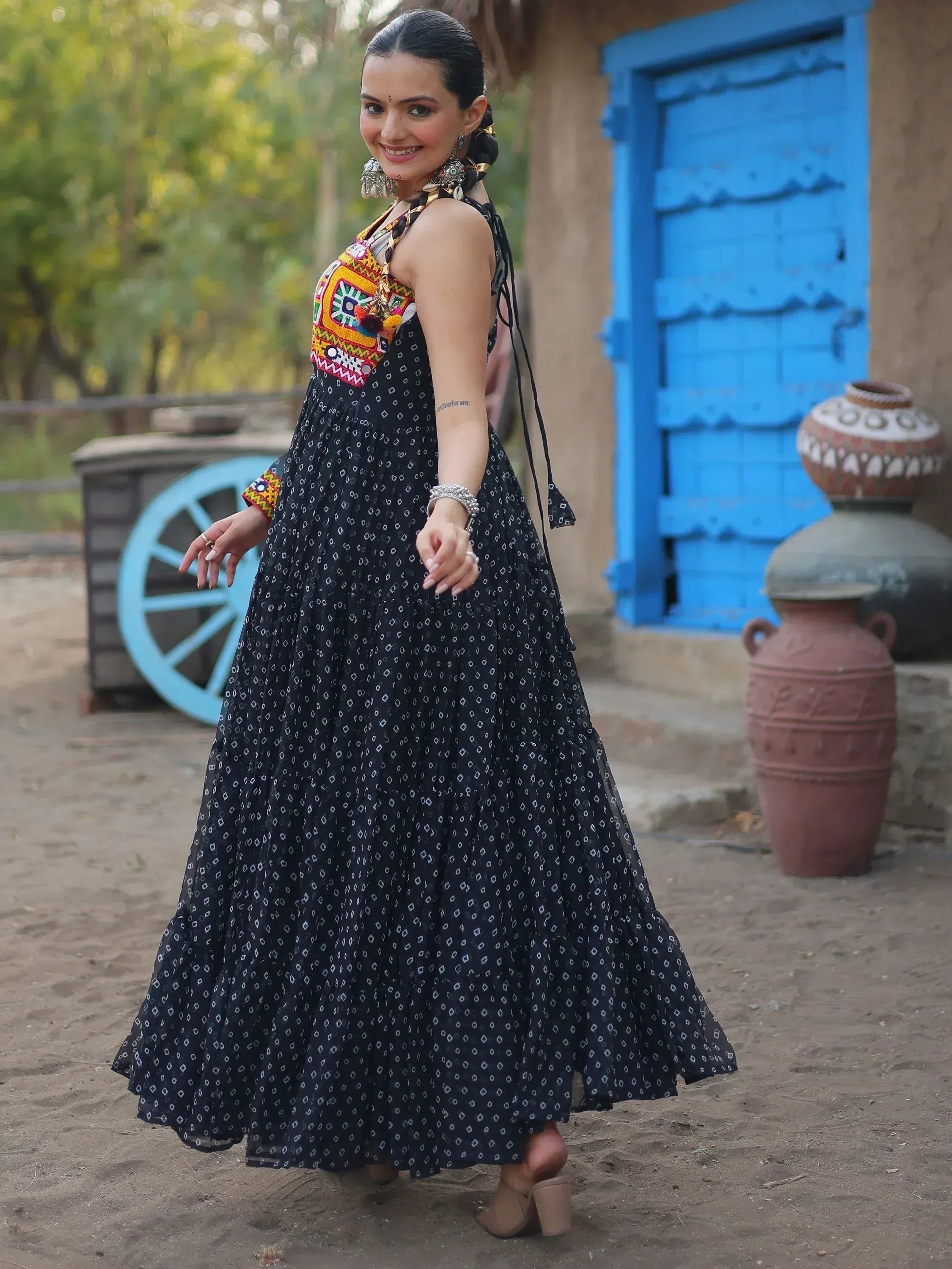 Woman in black ethnic gown with colorful embroidery, standing near blue door and clay pots