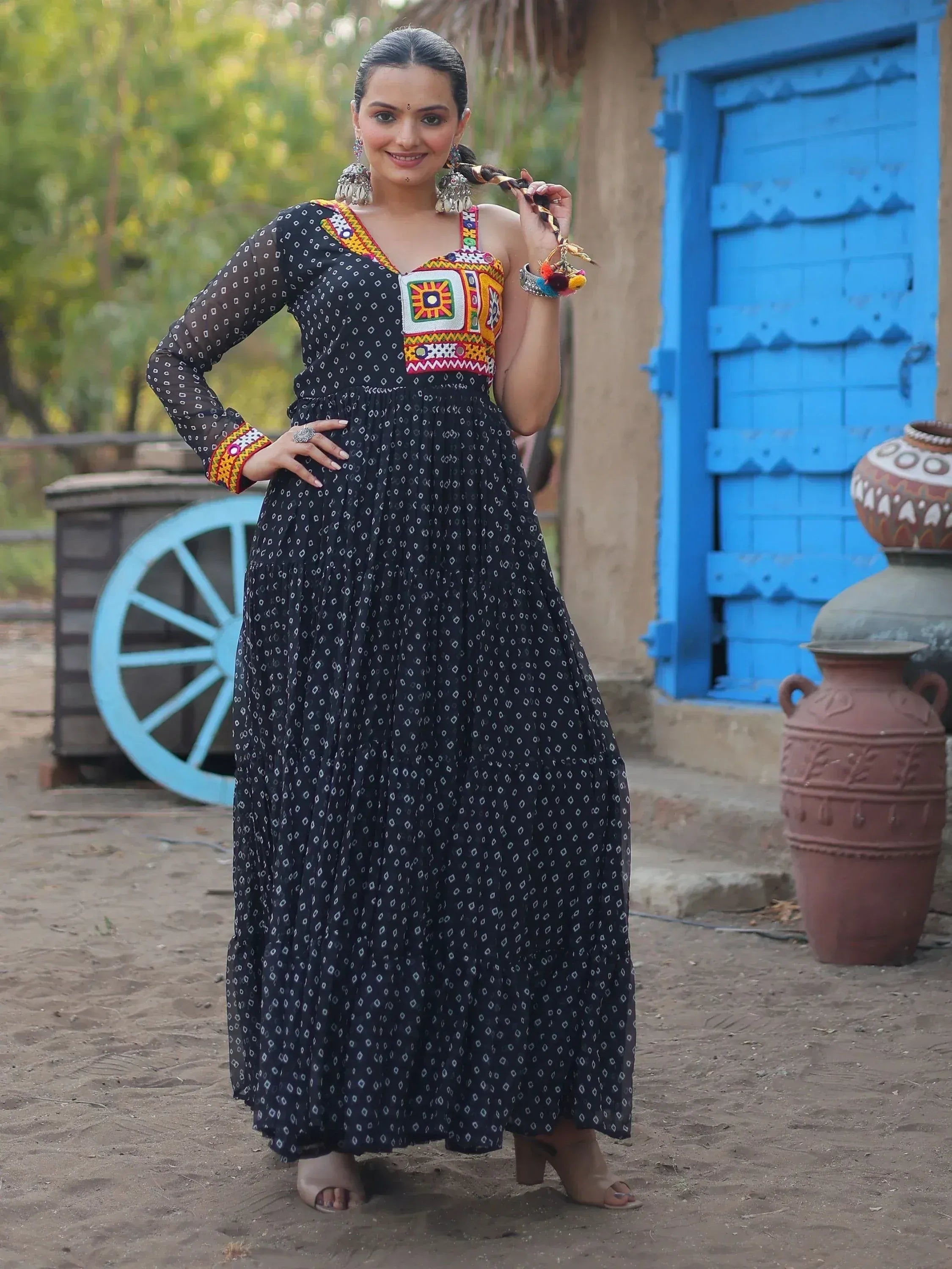 Woman in black ethnic gown with colorful embroidery, standing near blue door and clay pots.
