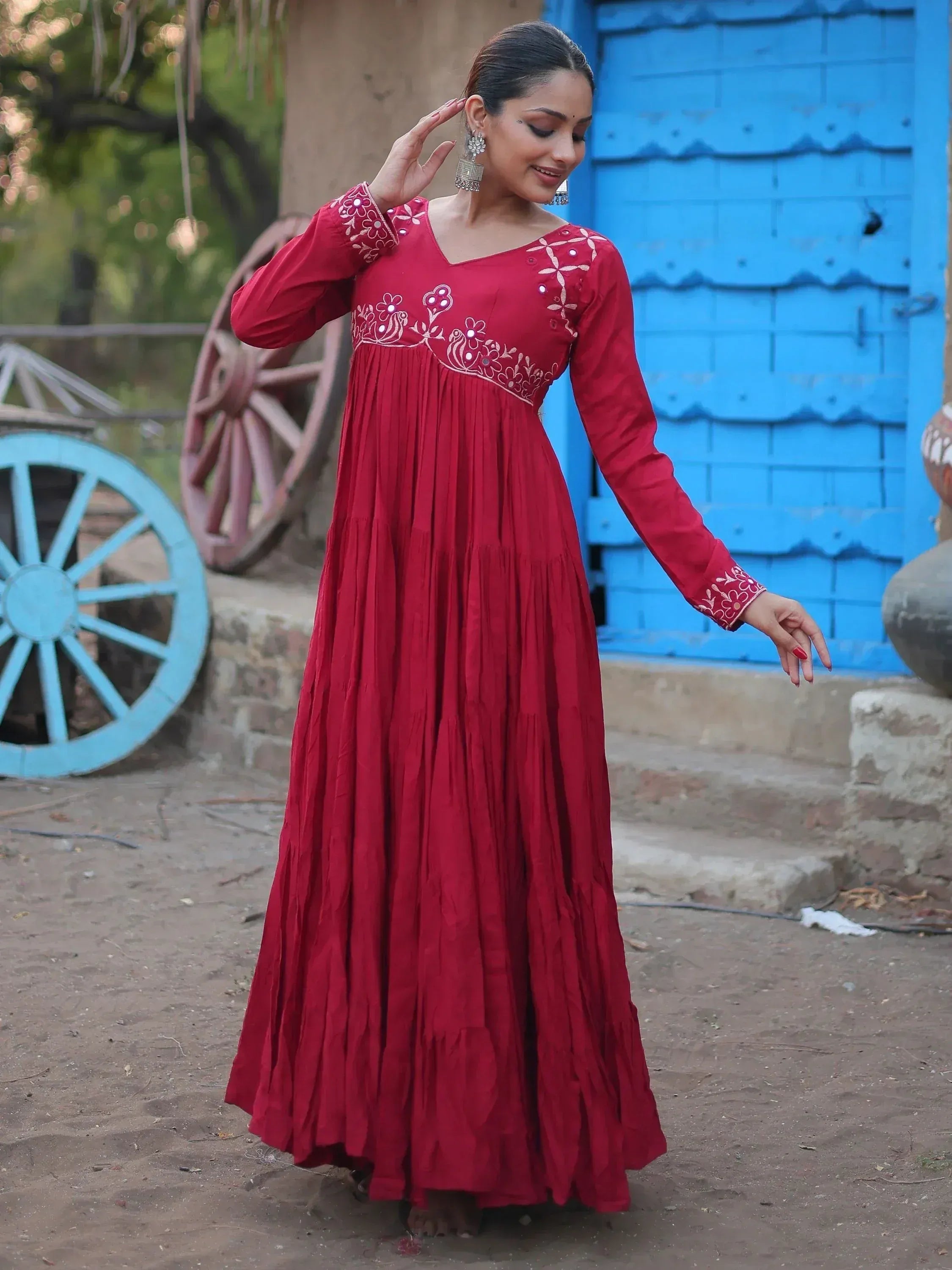 Red ethnic gown with floral embroidery, long sleeves, and pleated skirt, styled with jhumka earrings against a rustic blue door backdrop.