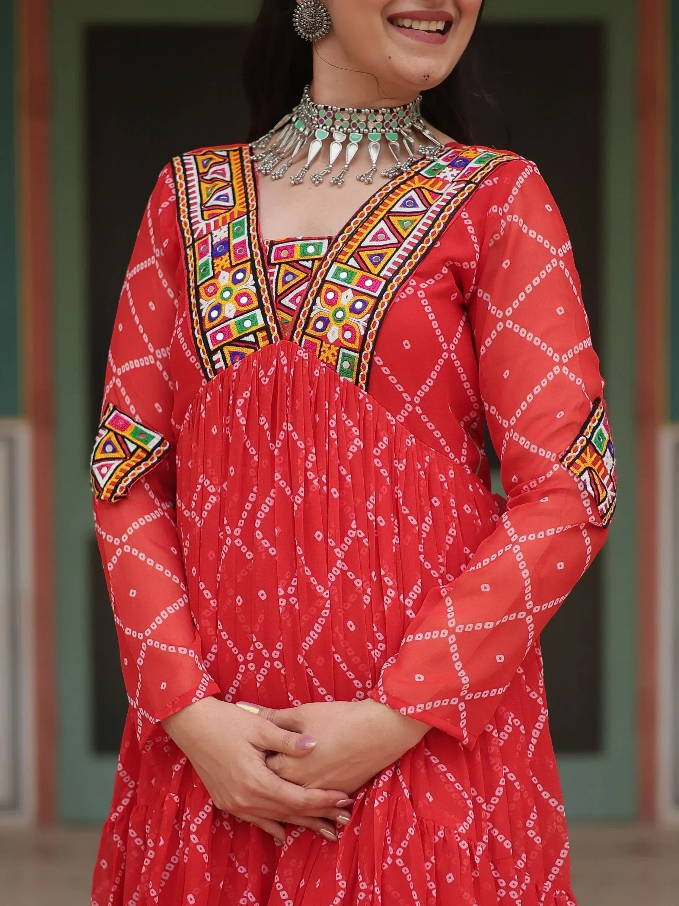 Red ethnic gown with colorful mirror work and geometric embroidery, paired with traditional silver jewelry.