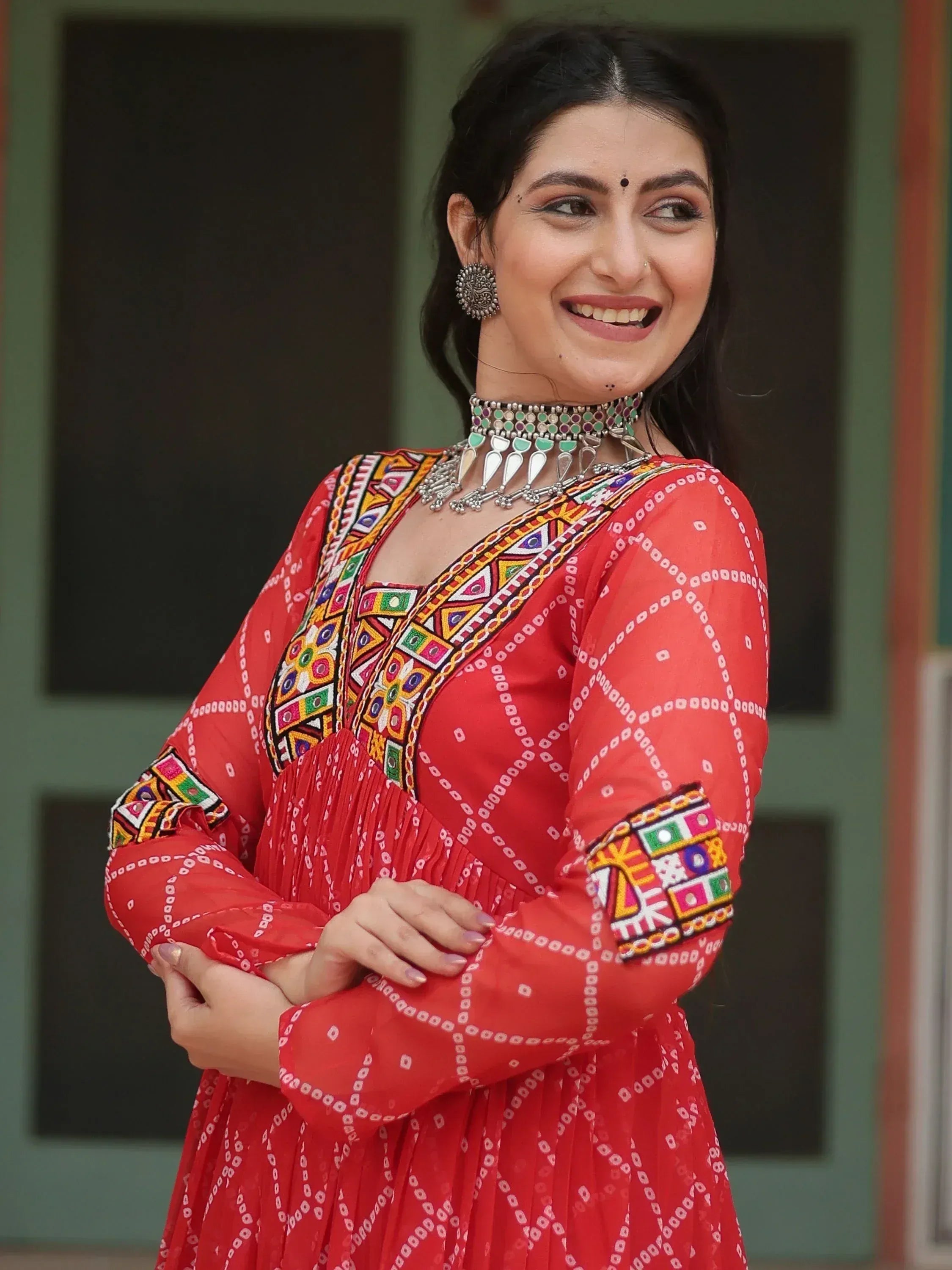 Woman in vibrant red ethnic gown with colorful mirror work and traditional jewelry, perfect for festive occasions.
