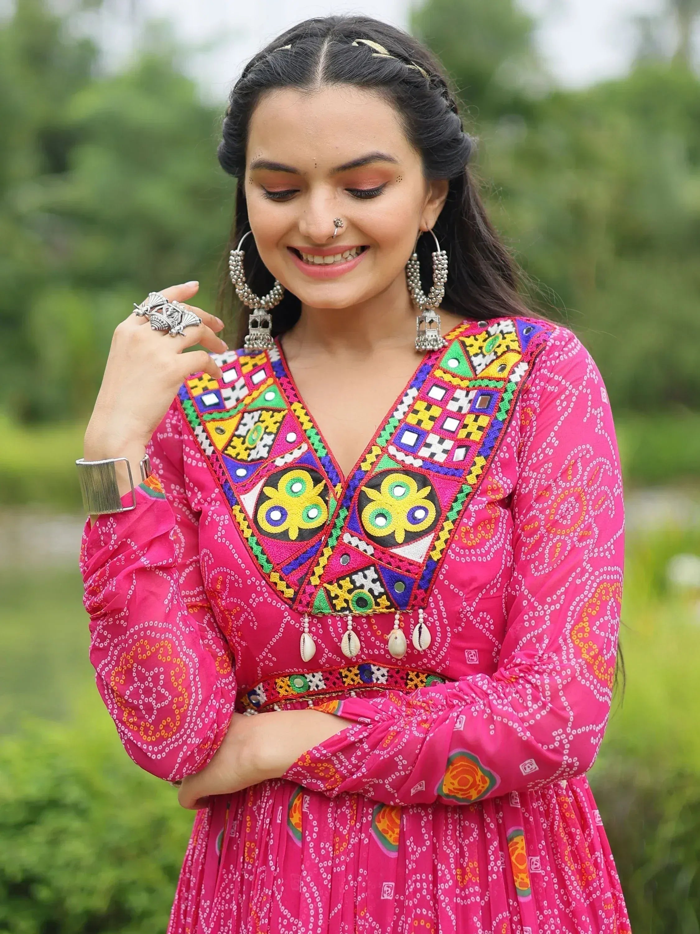 Woman in vibrant pink ethnic gown with colorful mirror work and traditional jewelry, perfect for festive occasions.
