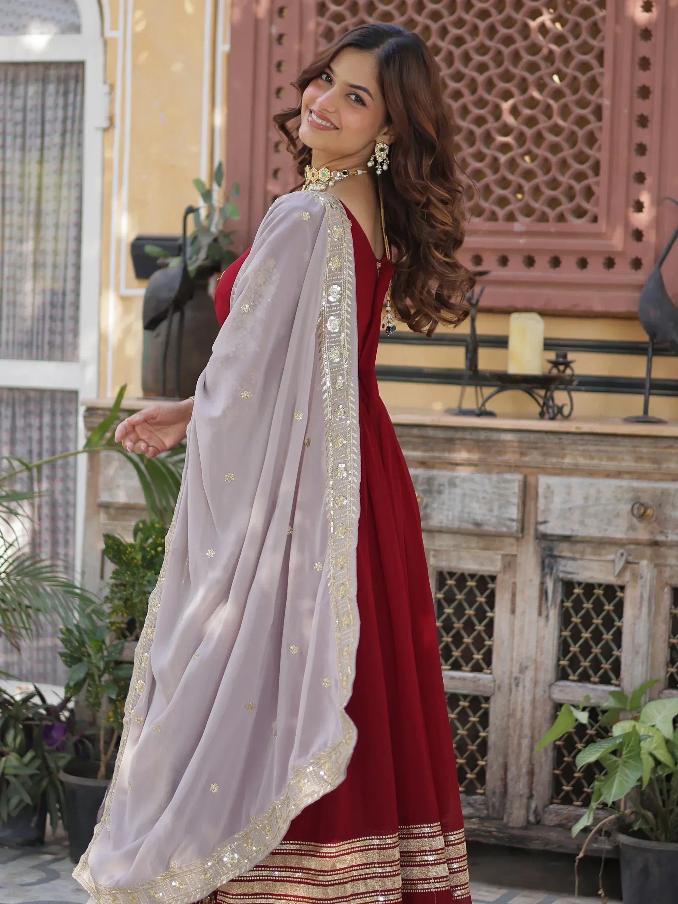 Woman in maroon ethnic gown with embroidered dupatta, floral jewelry, outdoor setting