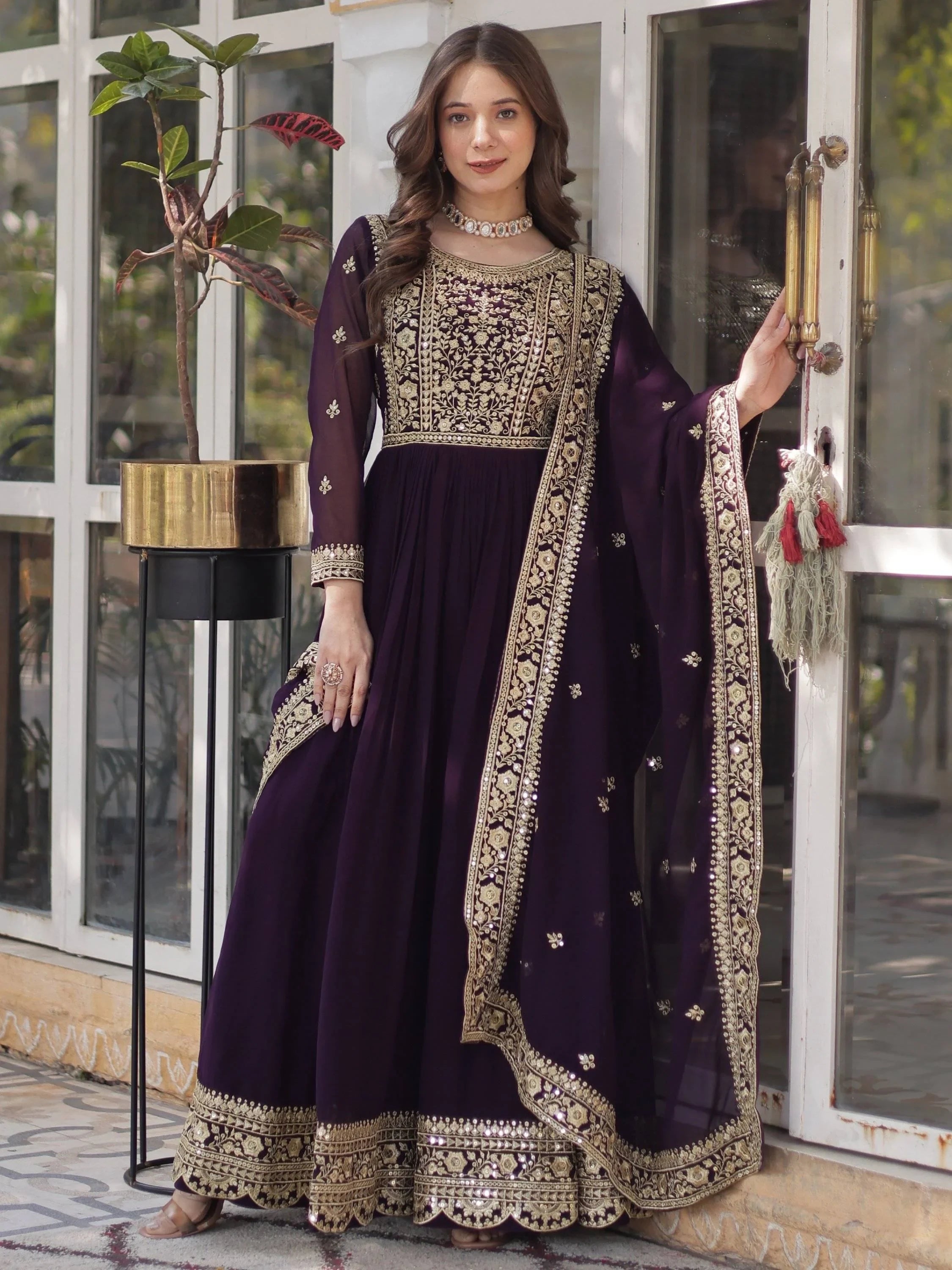 Woman wearing a wine-colored embroidered Anarkali gown with gold detailing, standing indoors