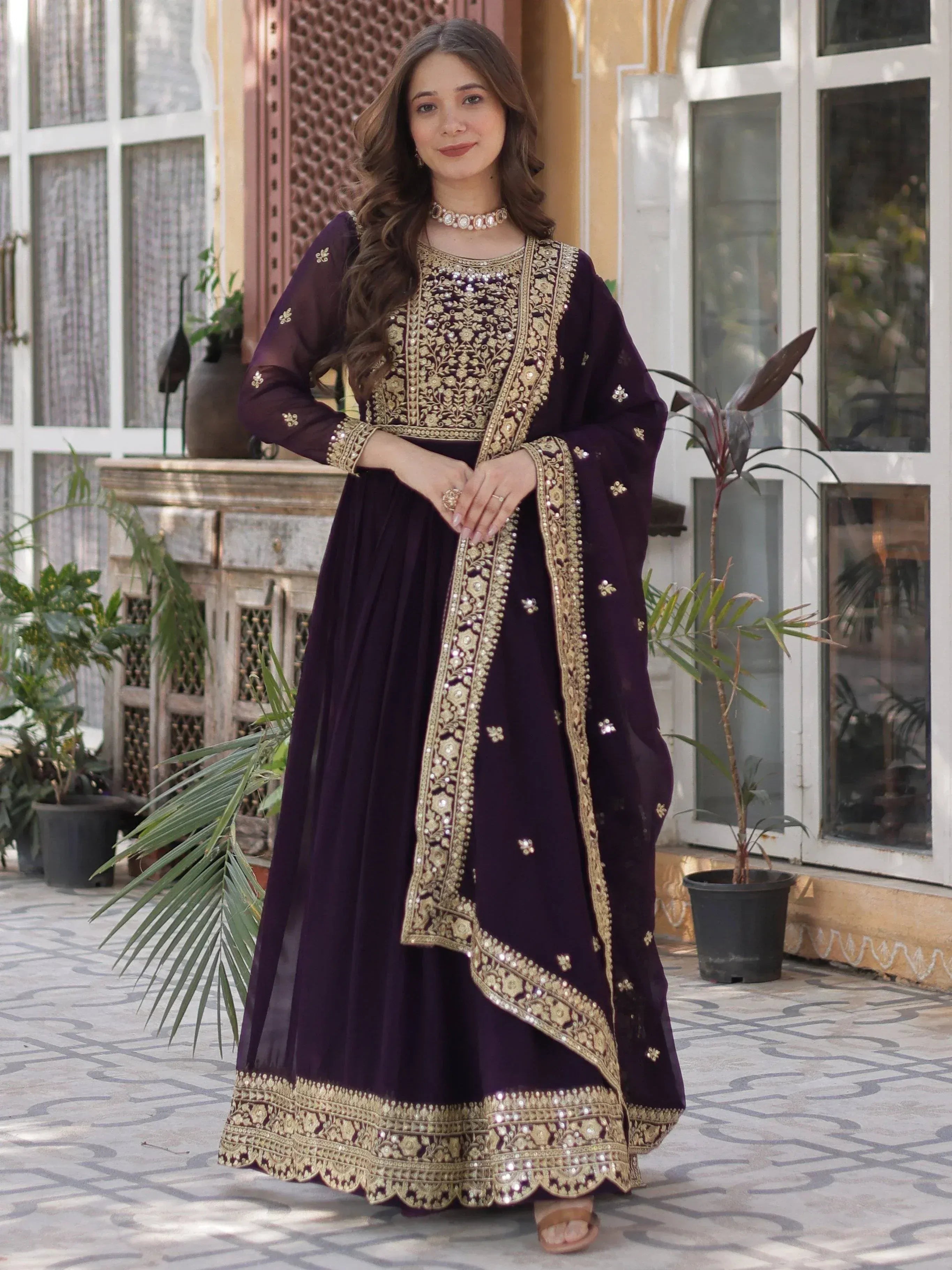Woman wearing a purple embroidered Anarkali gown with gold detailing, standing outdoors
