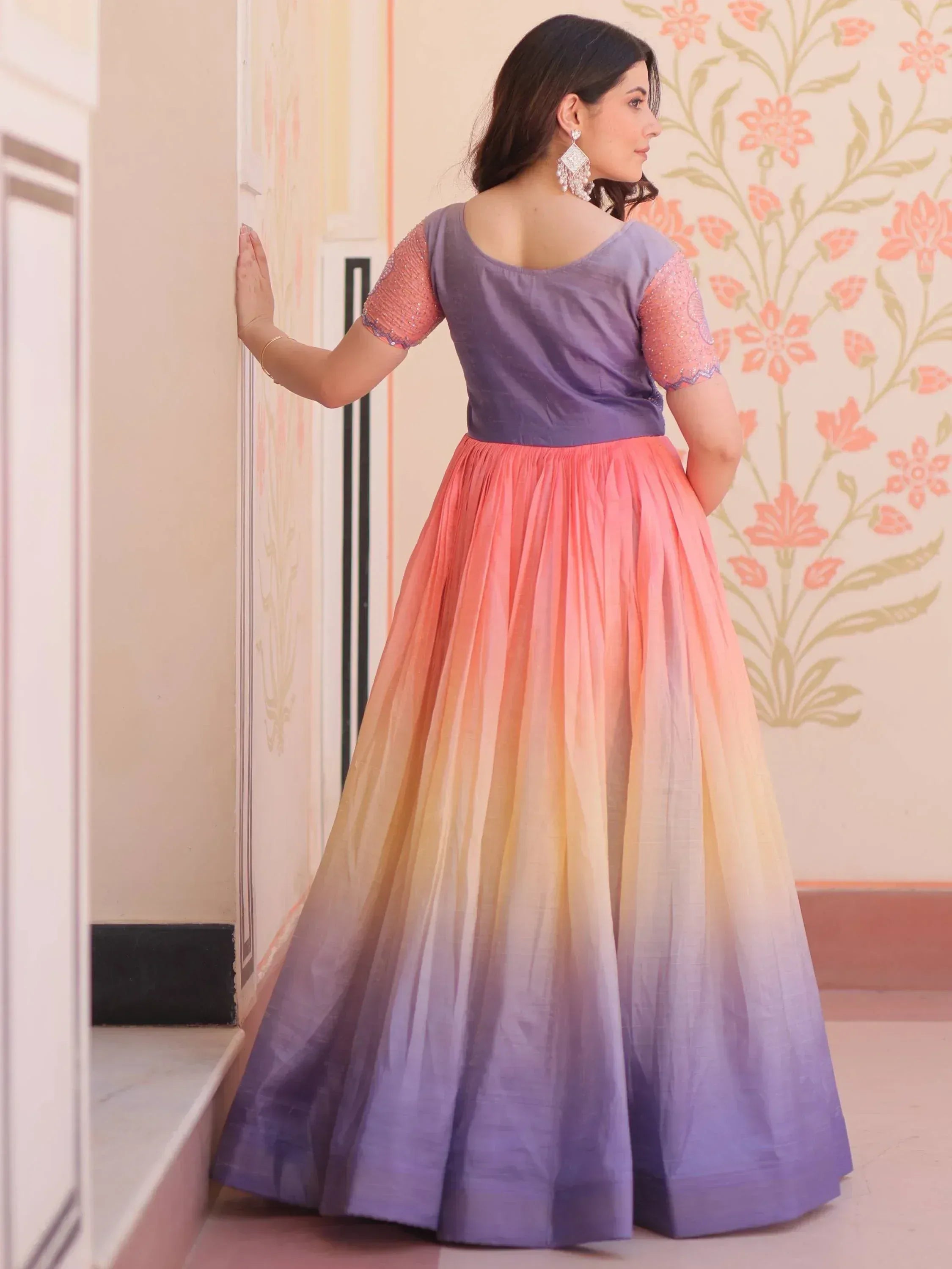 Woman in purple top and ombre pink-to-lavender ethnic gown, floral wall backdrop