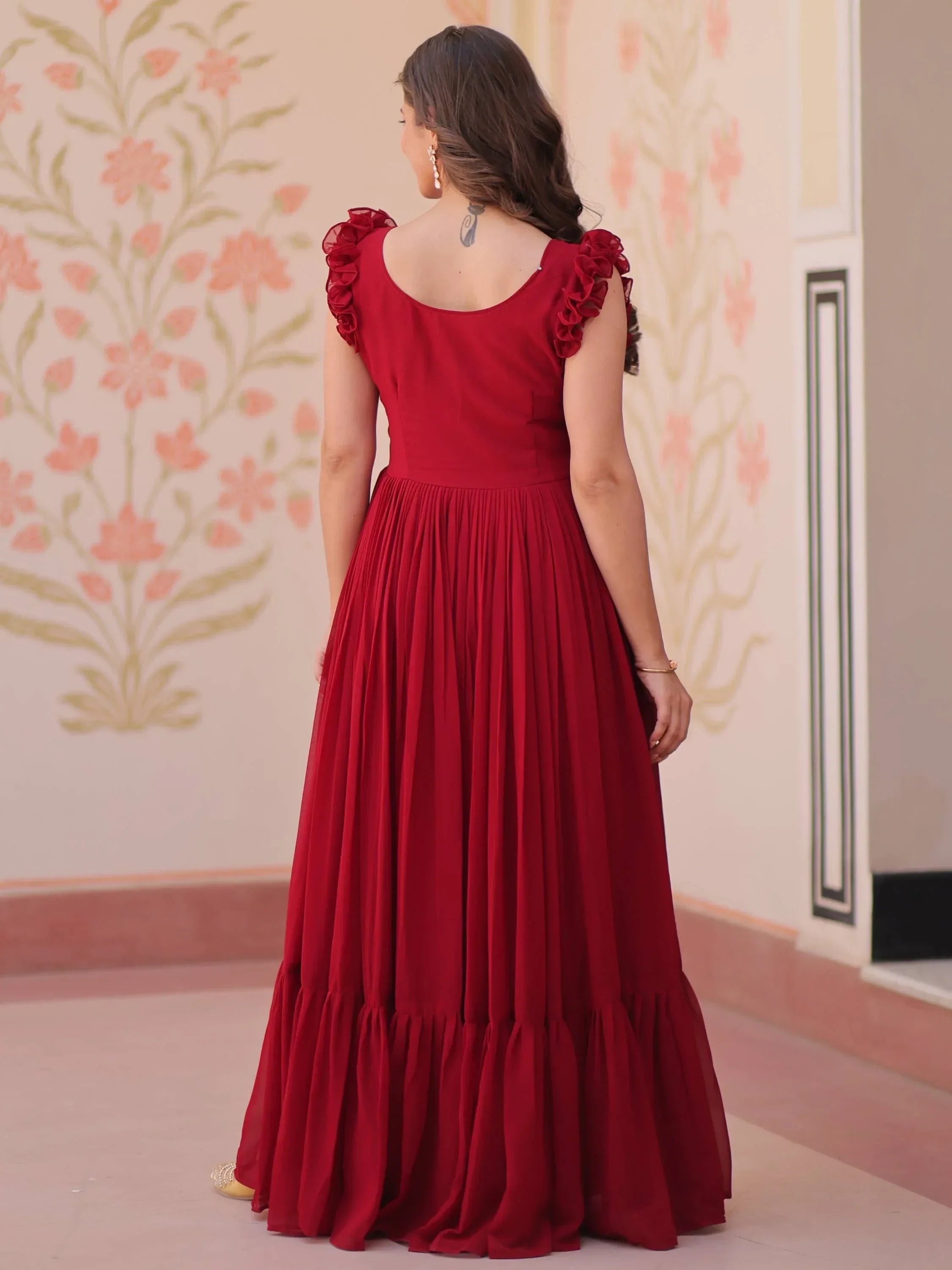 Woman in maroon ethnic gown with ruffled sleeves, standing indoors with floral wall art