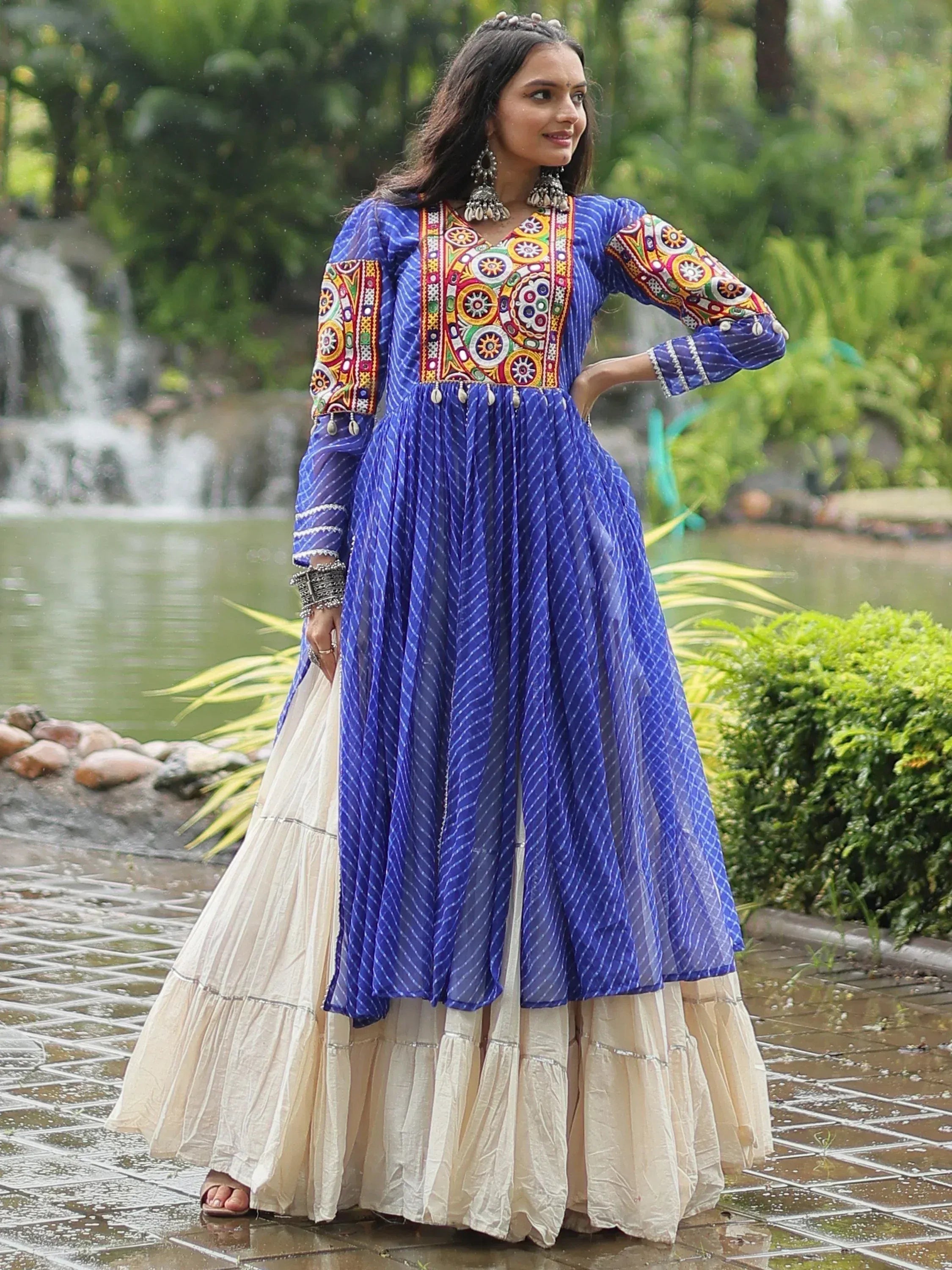 Blue embroidered ethnic gown with colorful patterns, paired with cream skirt, worn outdoors near a waterfall.