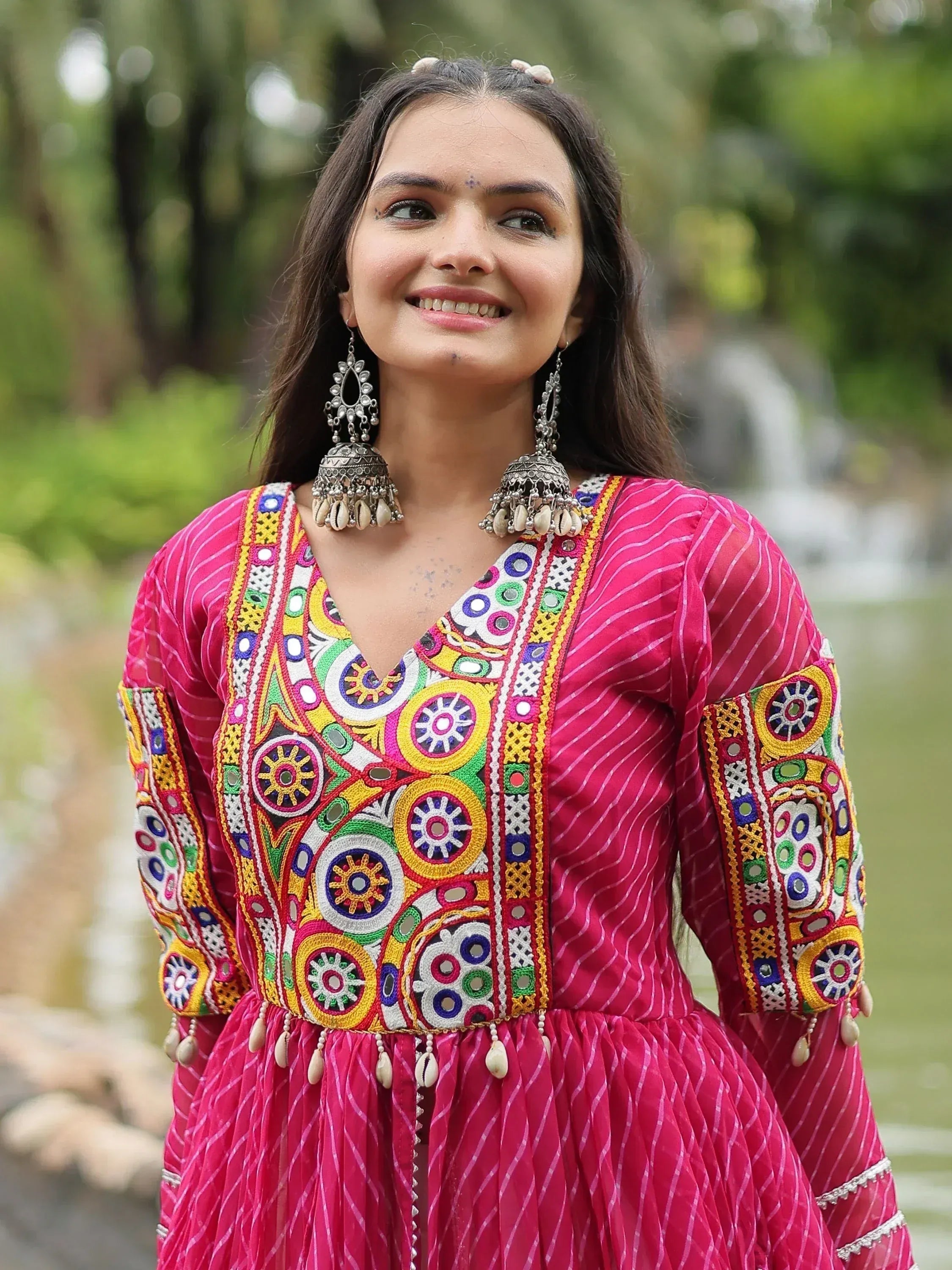 Woman in vibrant pink ethnic gown with colorful mirror embroidery and jhumka earrings, perfect for festive occasions.