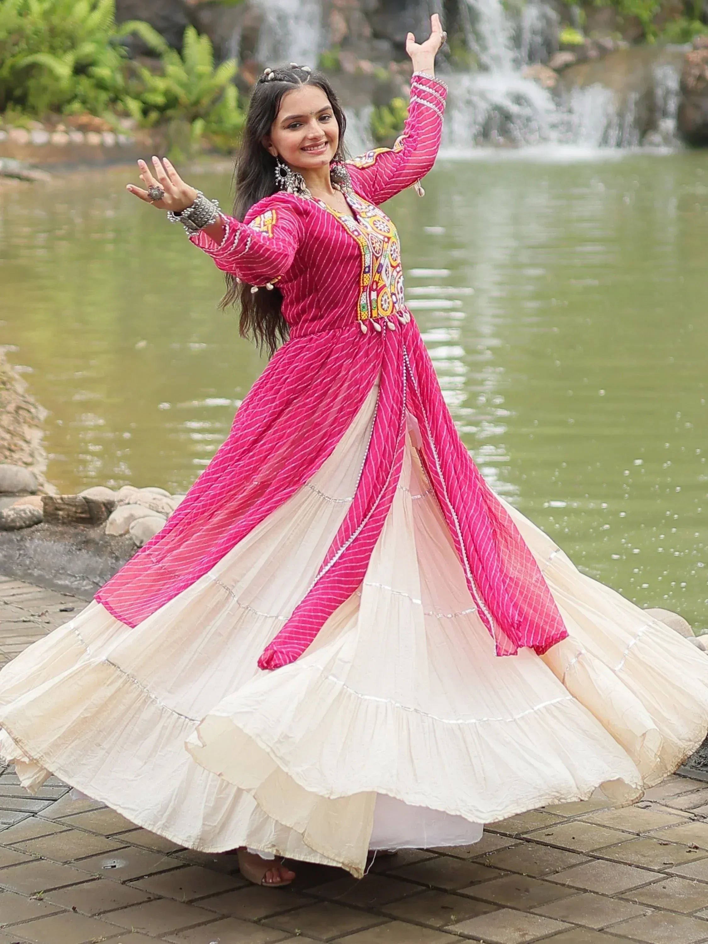 Woman in vibrant pink and cream ethnic gown dancing near waterfall, showcasing designer Indian wear