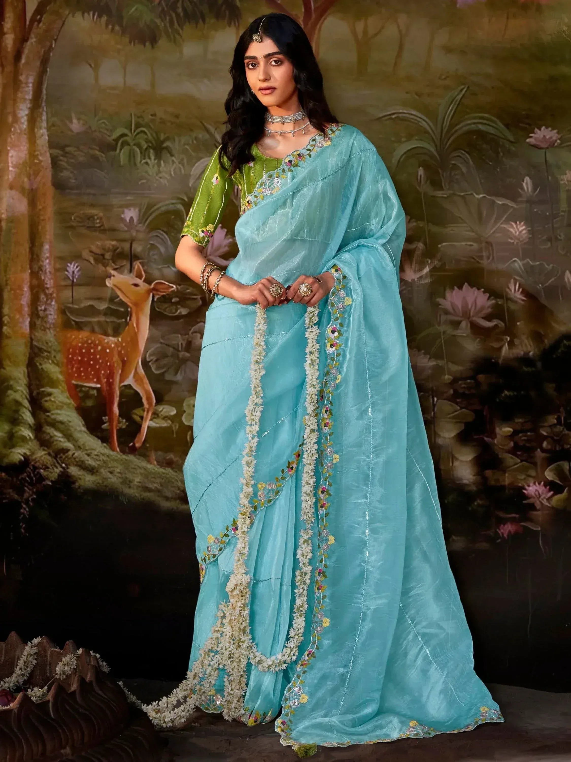 Woman in turquoise saree with floral embroidery and green blouse, styled with traditional jewelry against nature backdrop