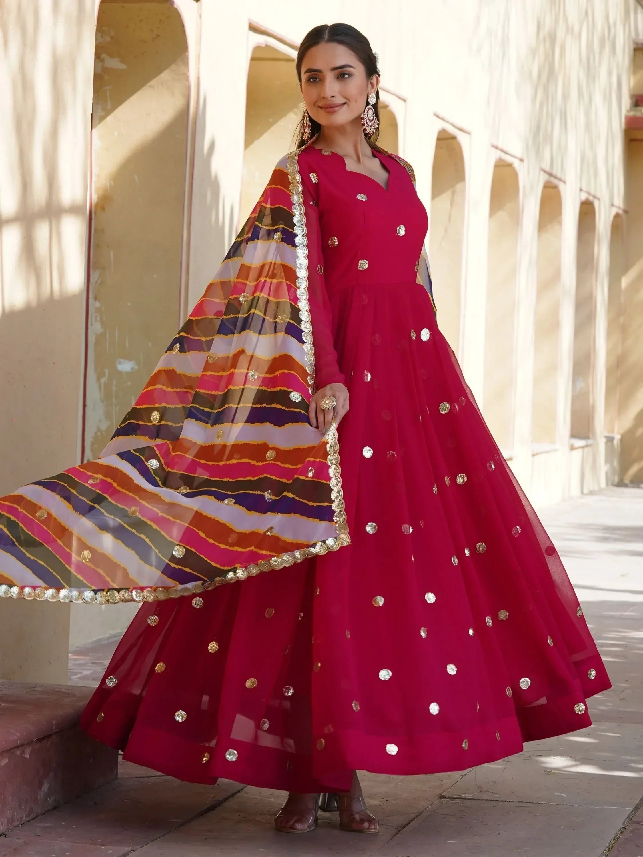 Woman in vibrant red Indian ethnic anarkali with gold mirror work and colorful striped dupatta, perfect for festive occasions.