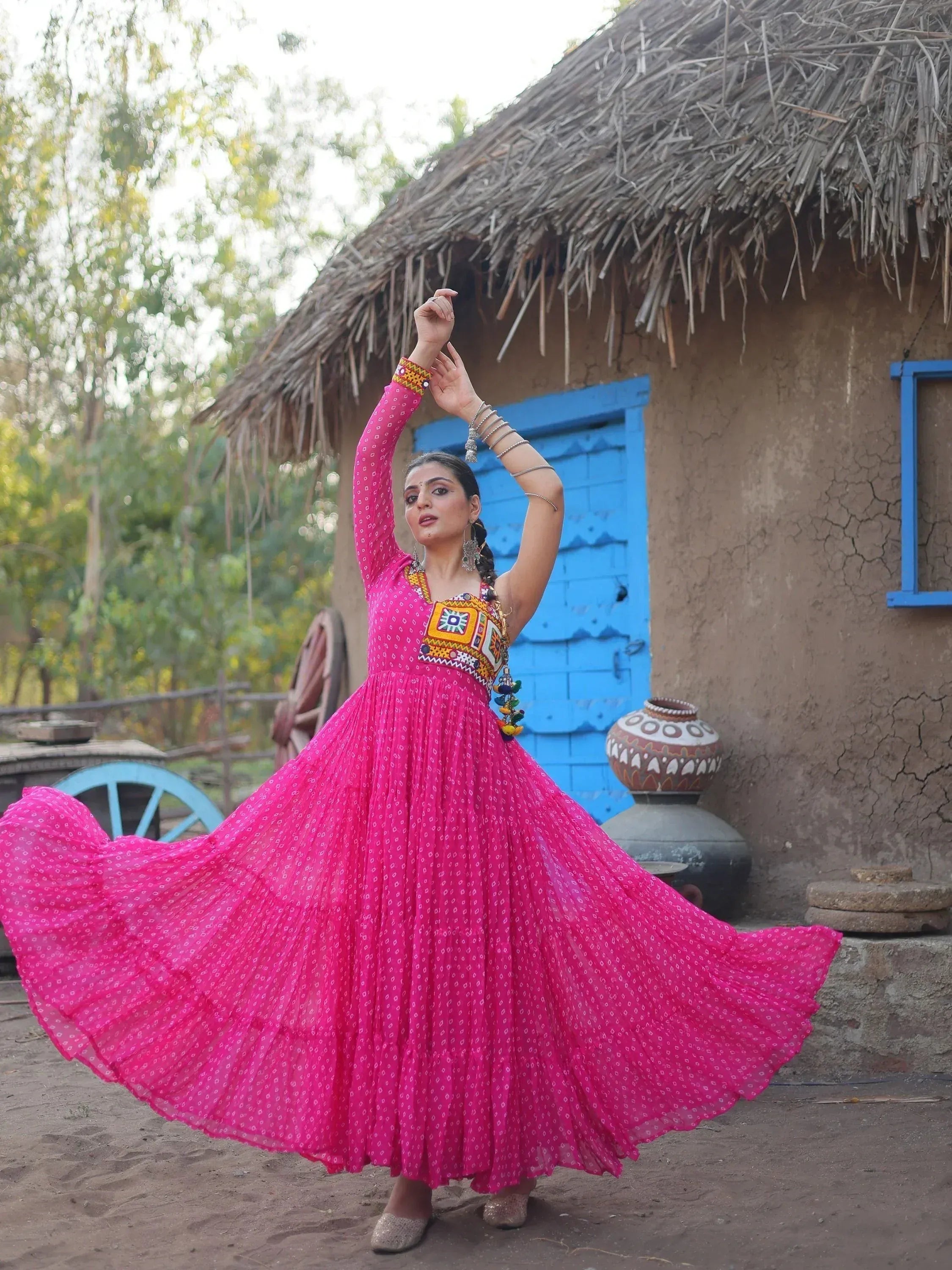 Woman in vibrant pink ethnic gown with mirror work, dancing near rustic hut, perfect for festive occasions.