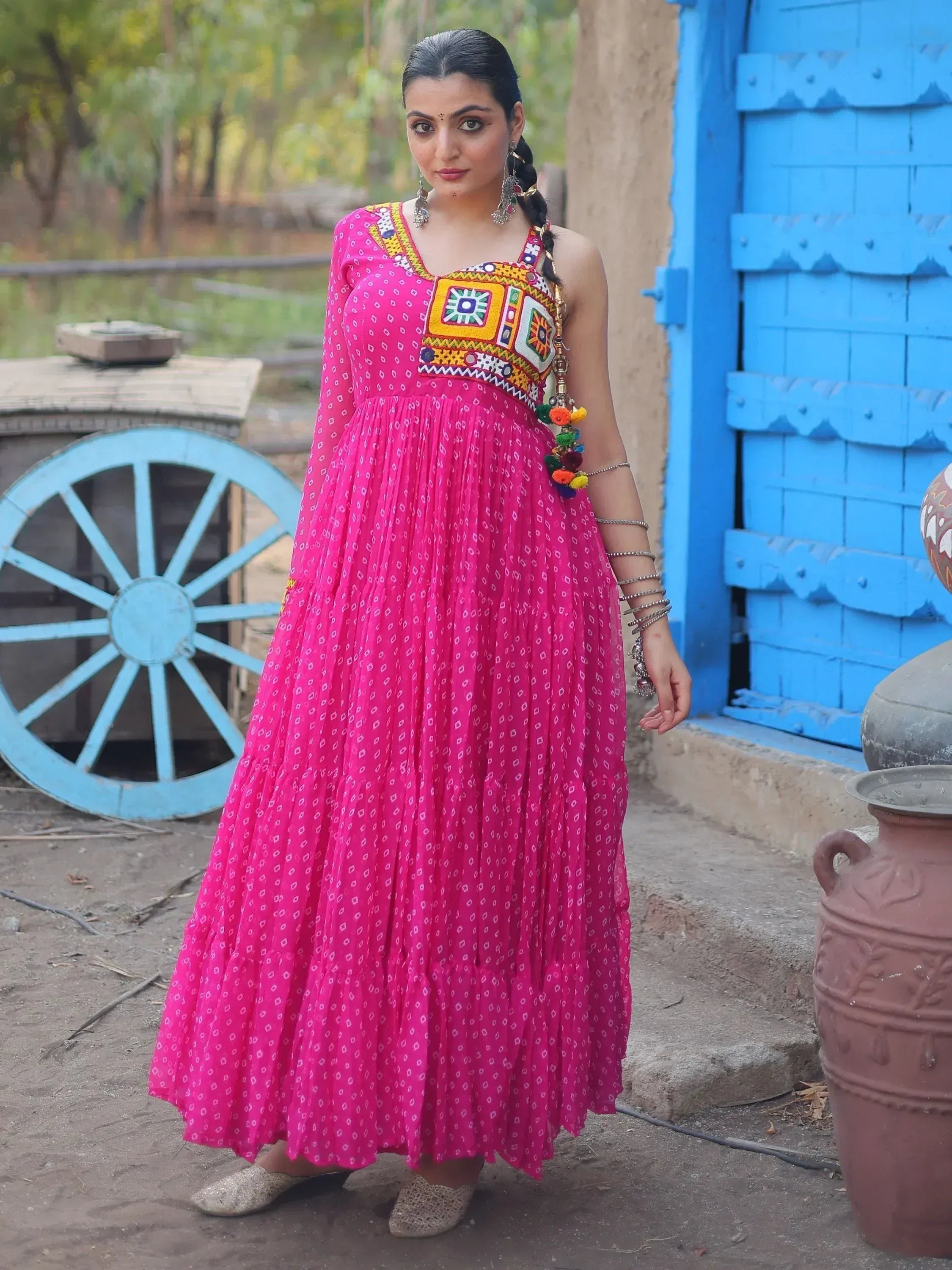 Woman in vibrant pink ethnic gown with mirror work, standing near blue door and cart wheel