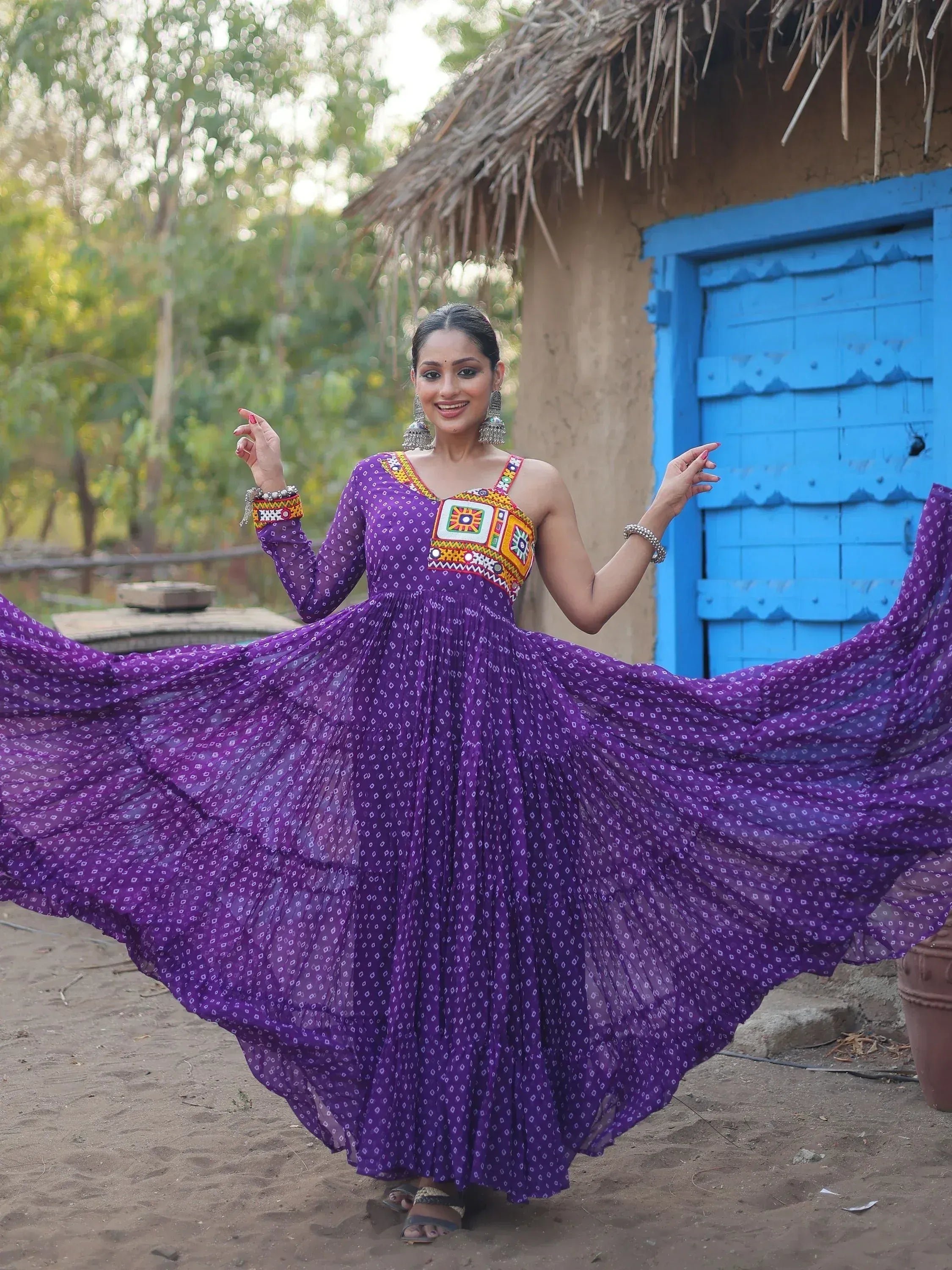 Woman in purple ethnic gown with mirror work, twirling near blue door and thatched hut