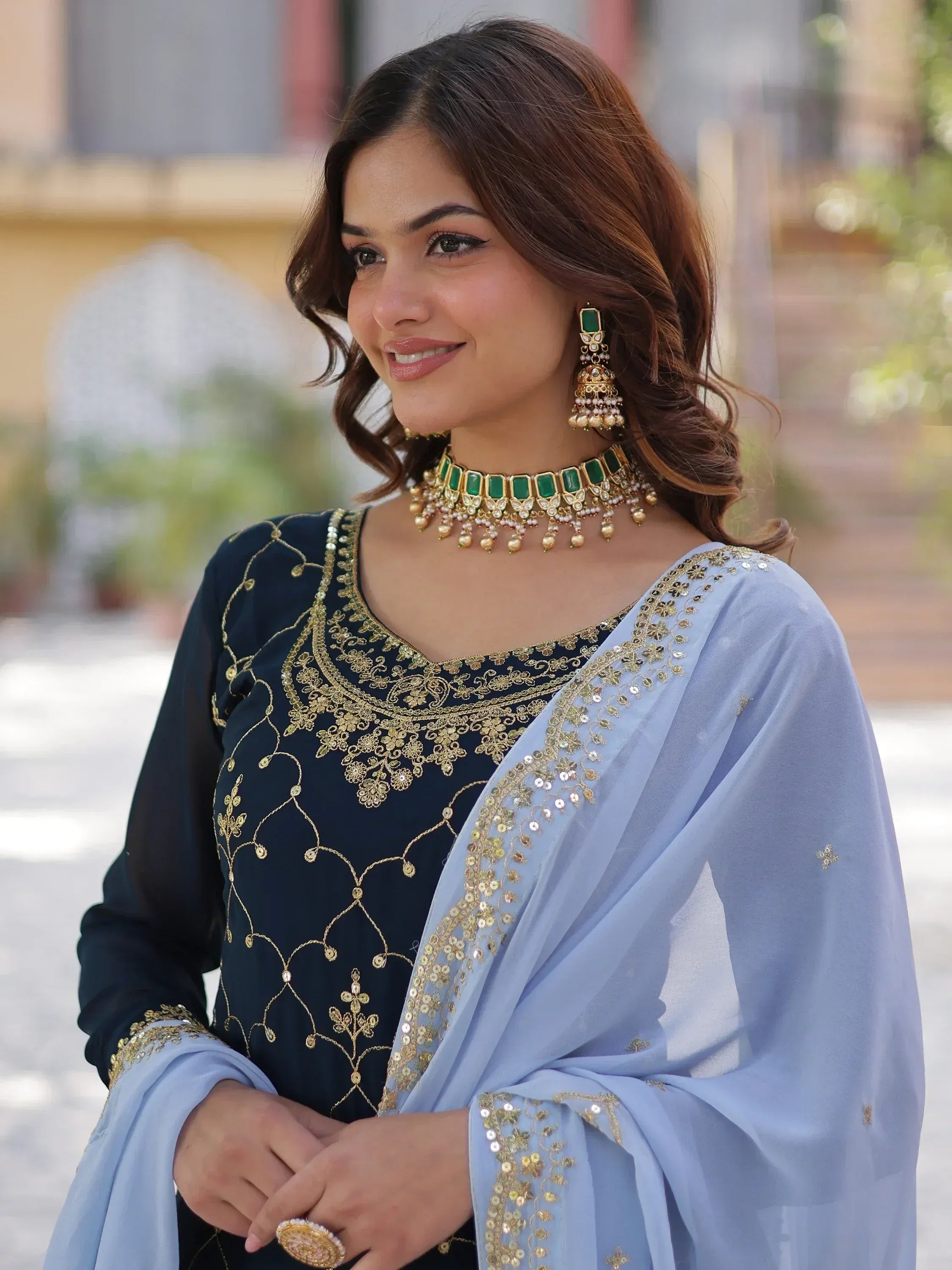 Elegant Indian ethnic wear with gold embroidery, light blue dupatta, and green gemstone jewelry