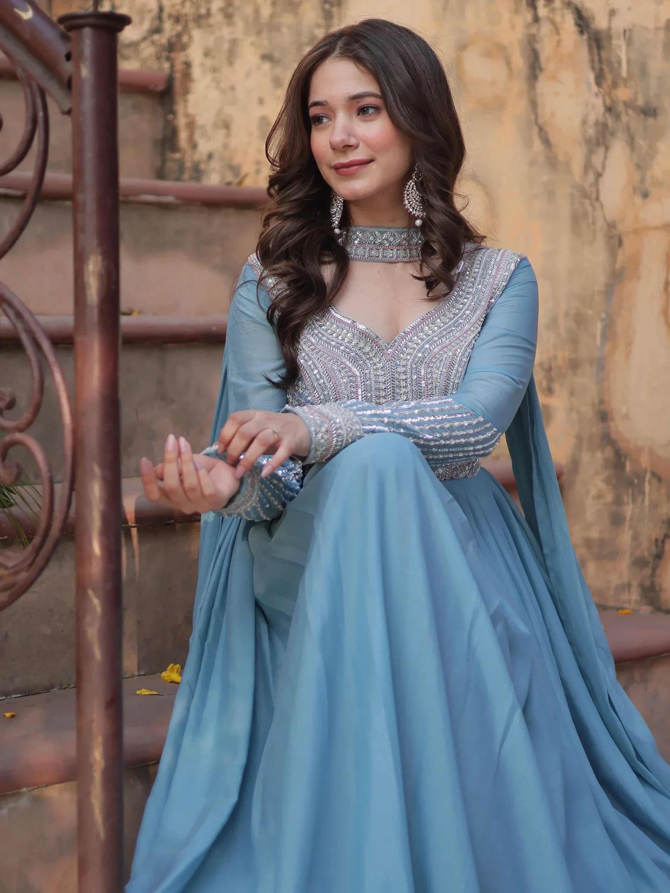 Woman in a sky blue embellished ethnic gown with dupatta, sitting on stairs outdoors.