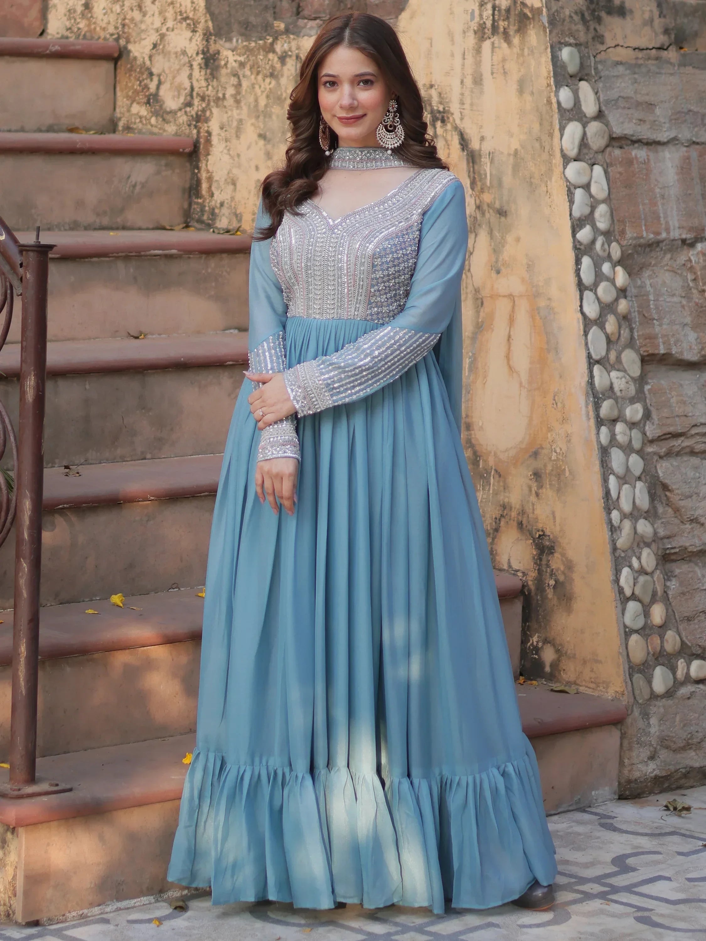 Woman in light blue embroidered ethnic gown with sheer neckline and long sleeves, standing outdoors.