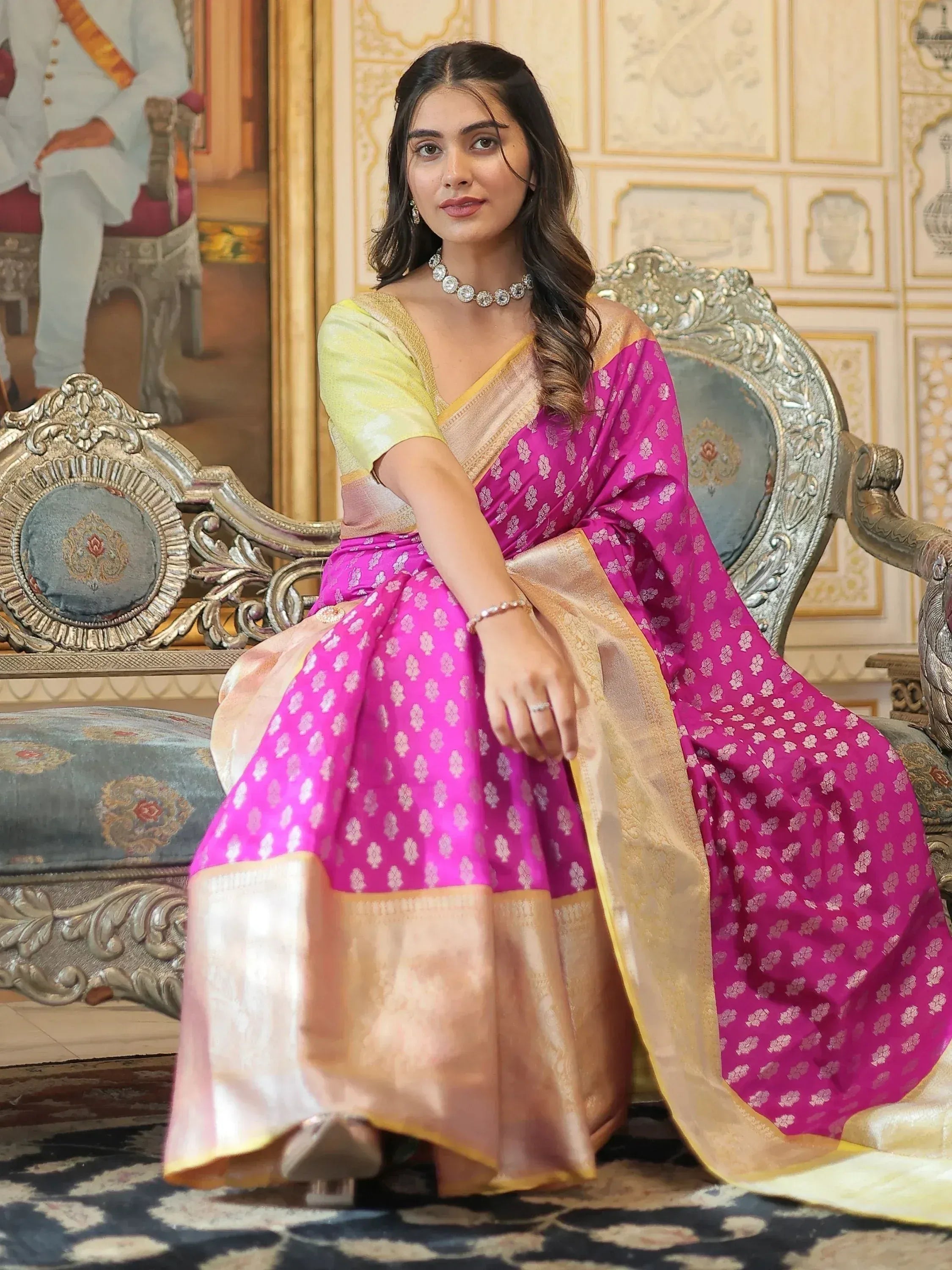 Woman in vibrant pink and gold ethnic saree with floral pattern, seated on ornate chair