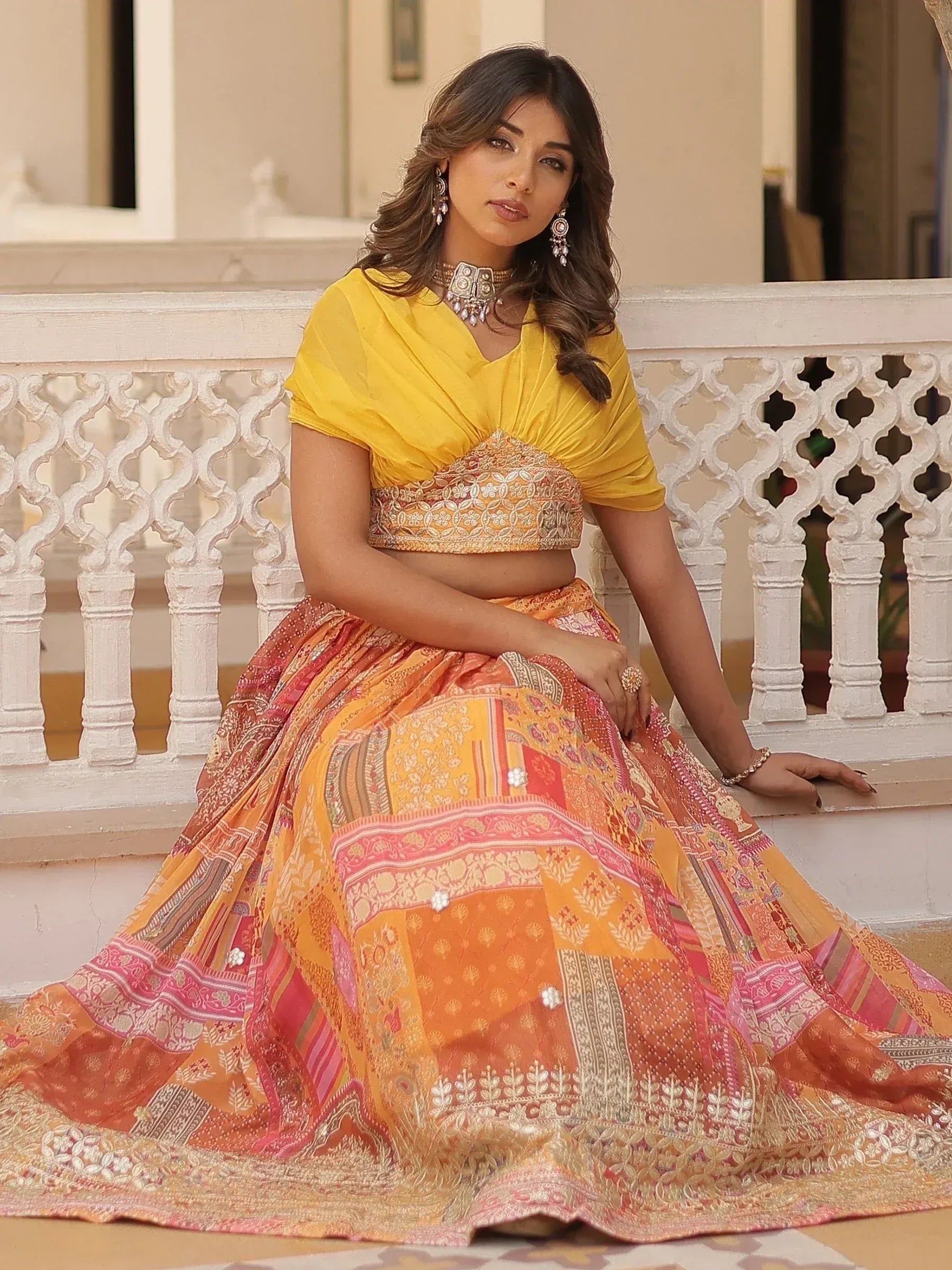 Woman in vibrant yellow and orange embroidered lehenga choli, traditional Indian ethnic wear, seated on ornate balcony.