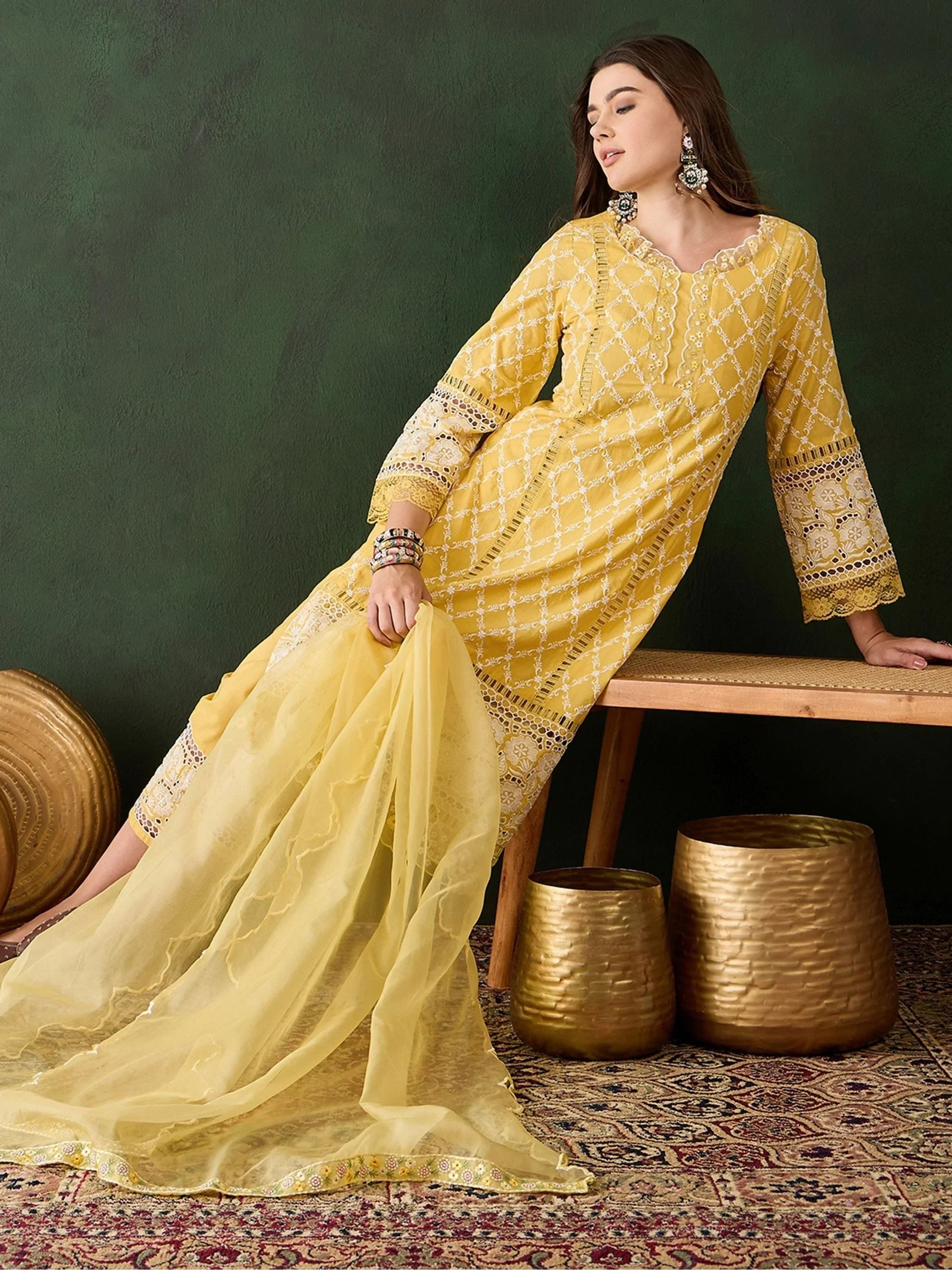 Woman in a yellow embroidered salwar kameez with dupatta, seated on a bench, ethnic decor.