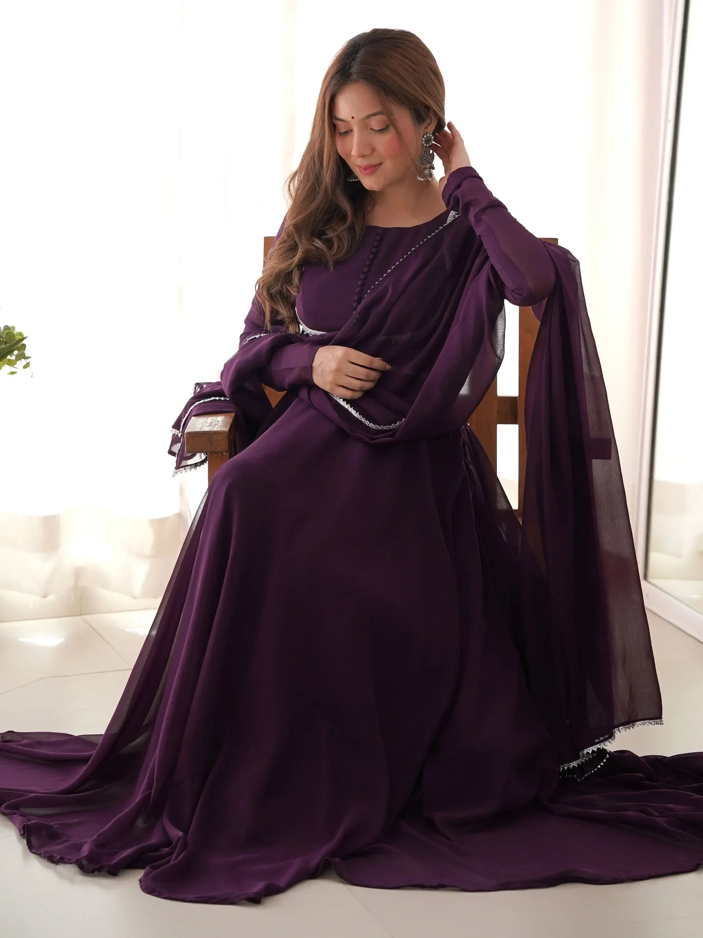 Woman in a flowing purple ethnic gown with dupatta, seated indoors, traditional attire