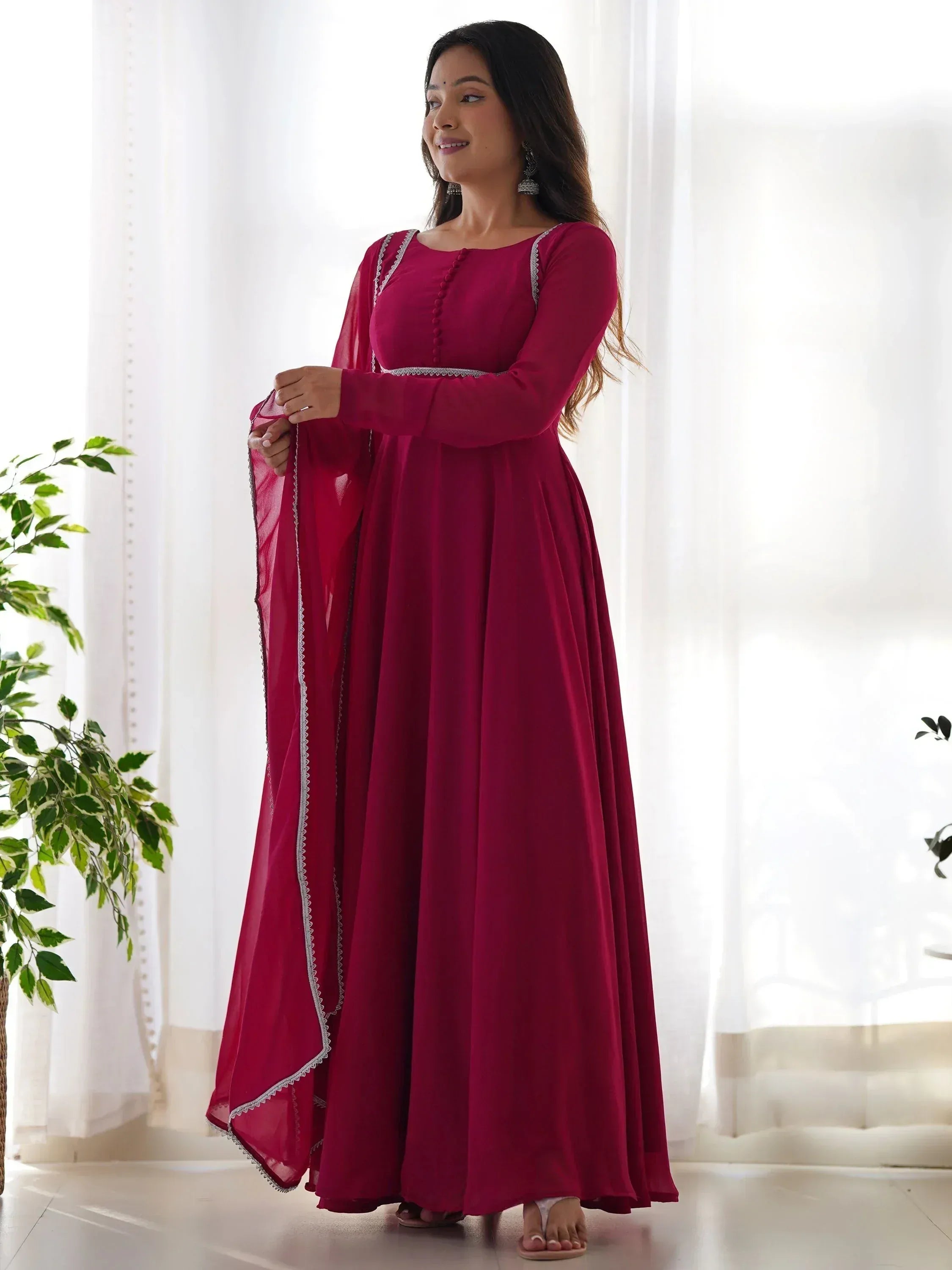 Woman in maroon ethnic gown with silver trim, long sleeves, and matching dupatta, standing near sheer curtains