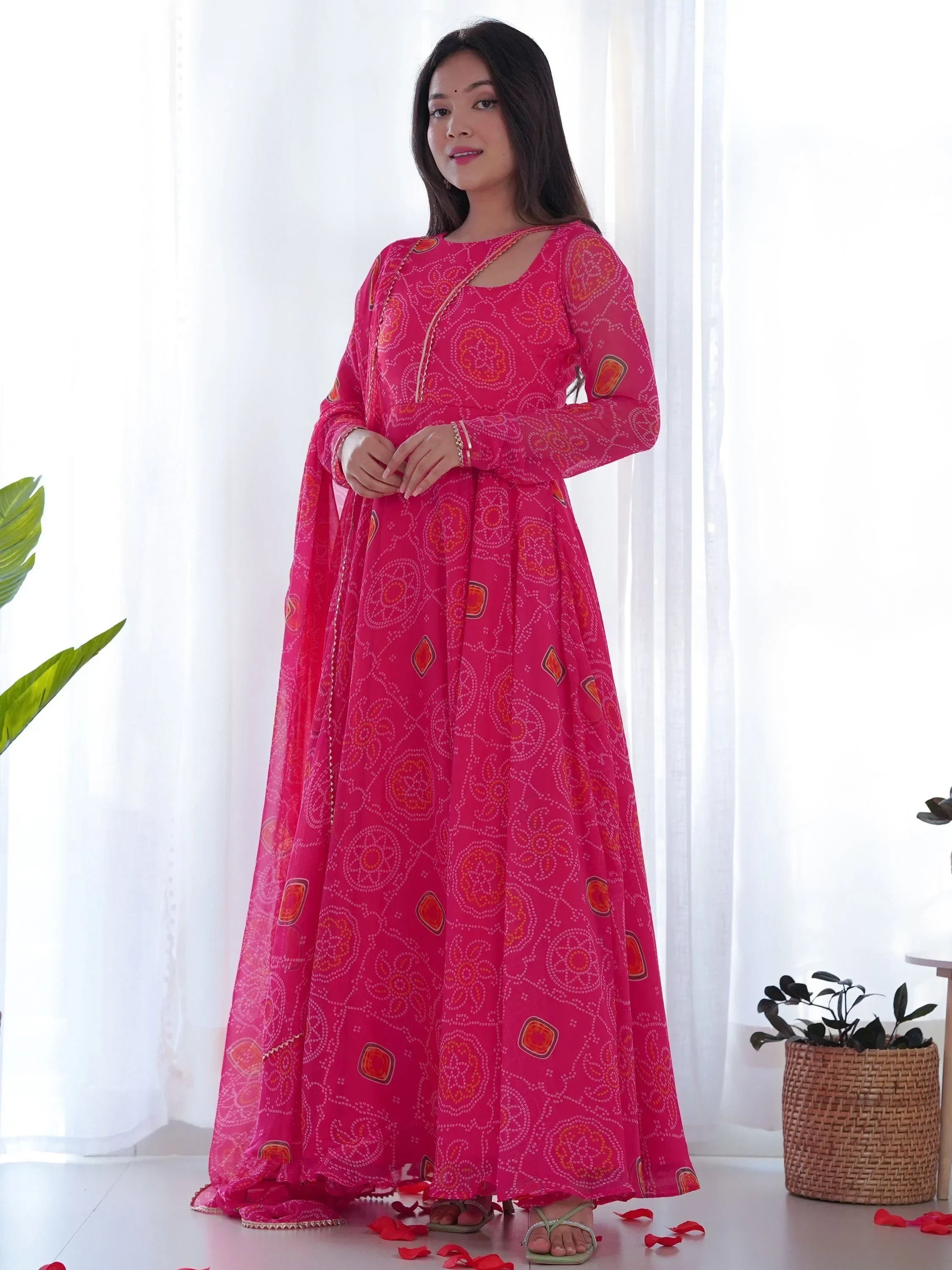 Woman in a bright pink bandhani print Indian ethnic gown with dupatta, standing indoors.