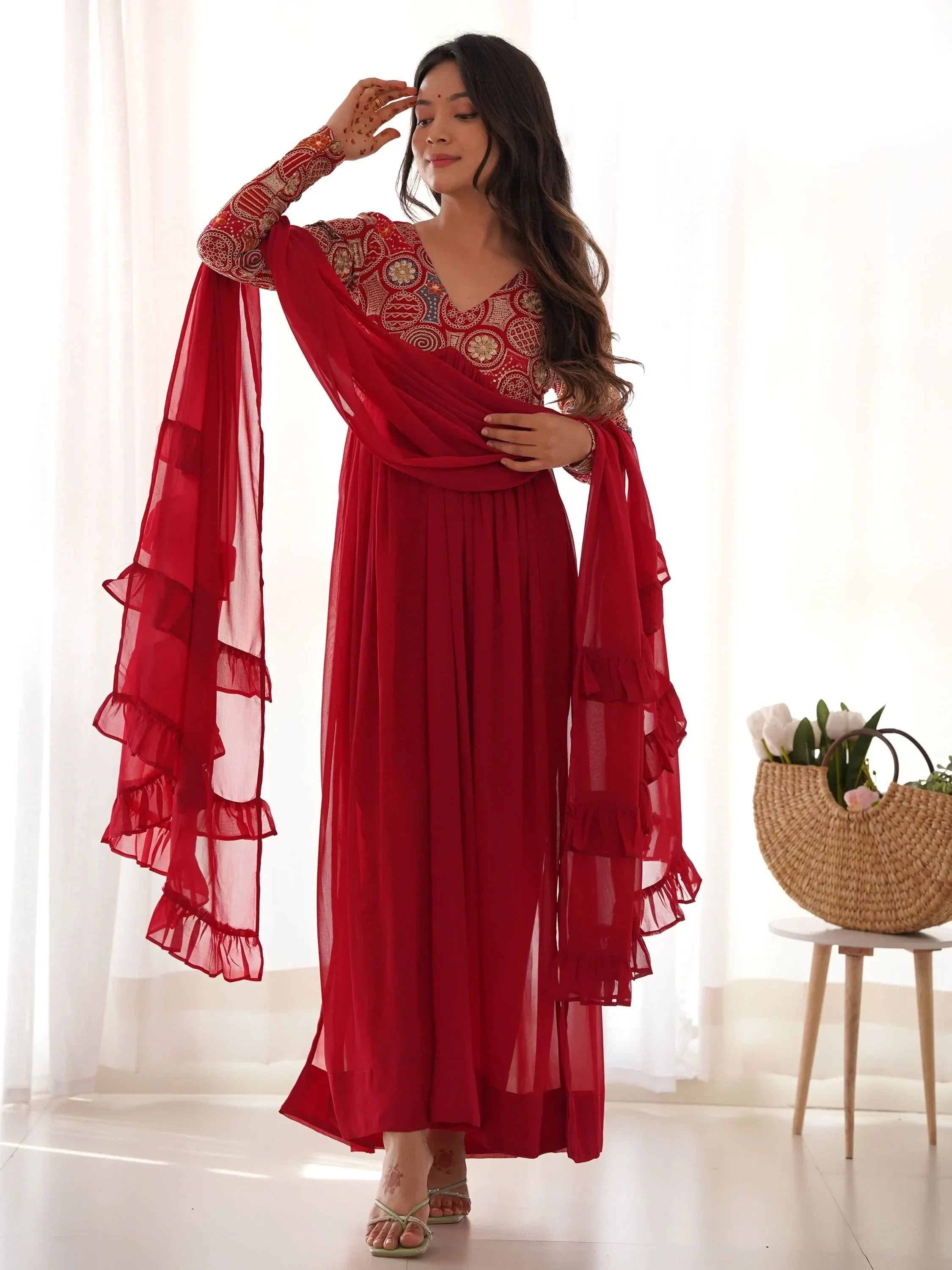 Woman in red embroidered ethnic gown with ruffled dupatta, standing indoors