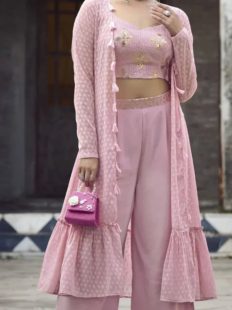 Woman in pink embroidered ethnic crop top, flared pants, and long jacket holding matching purse