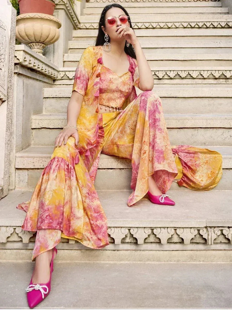 Woman in floral yellow and pink ethnic co-ord set with flared pants, statement earrings, and sunglasses, seated on ornate outdoor steps.