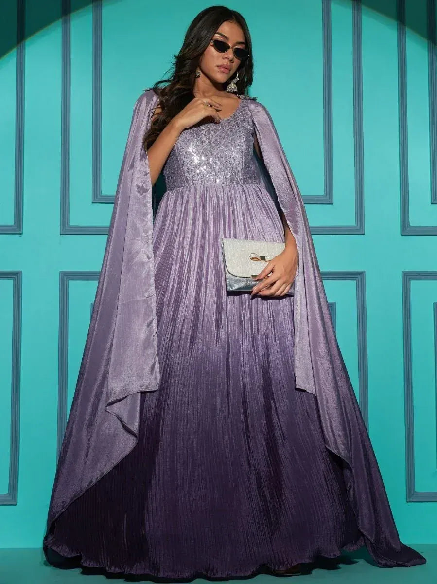 Woman in a lavender ethnic gown with cape sleeves, holding a clutch, posing against teal wall