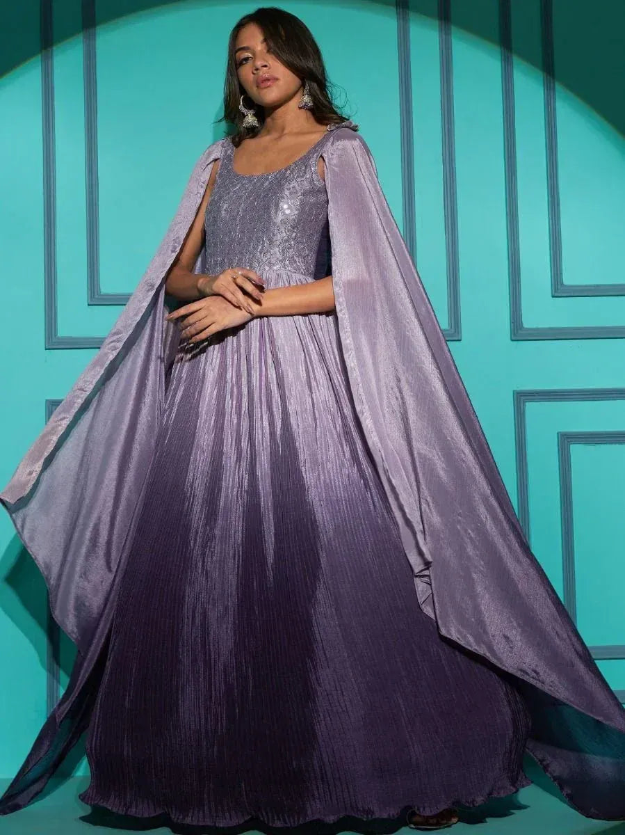 Woman in a lavender pleated ethnic gown with cape, standing against teal paneled wall